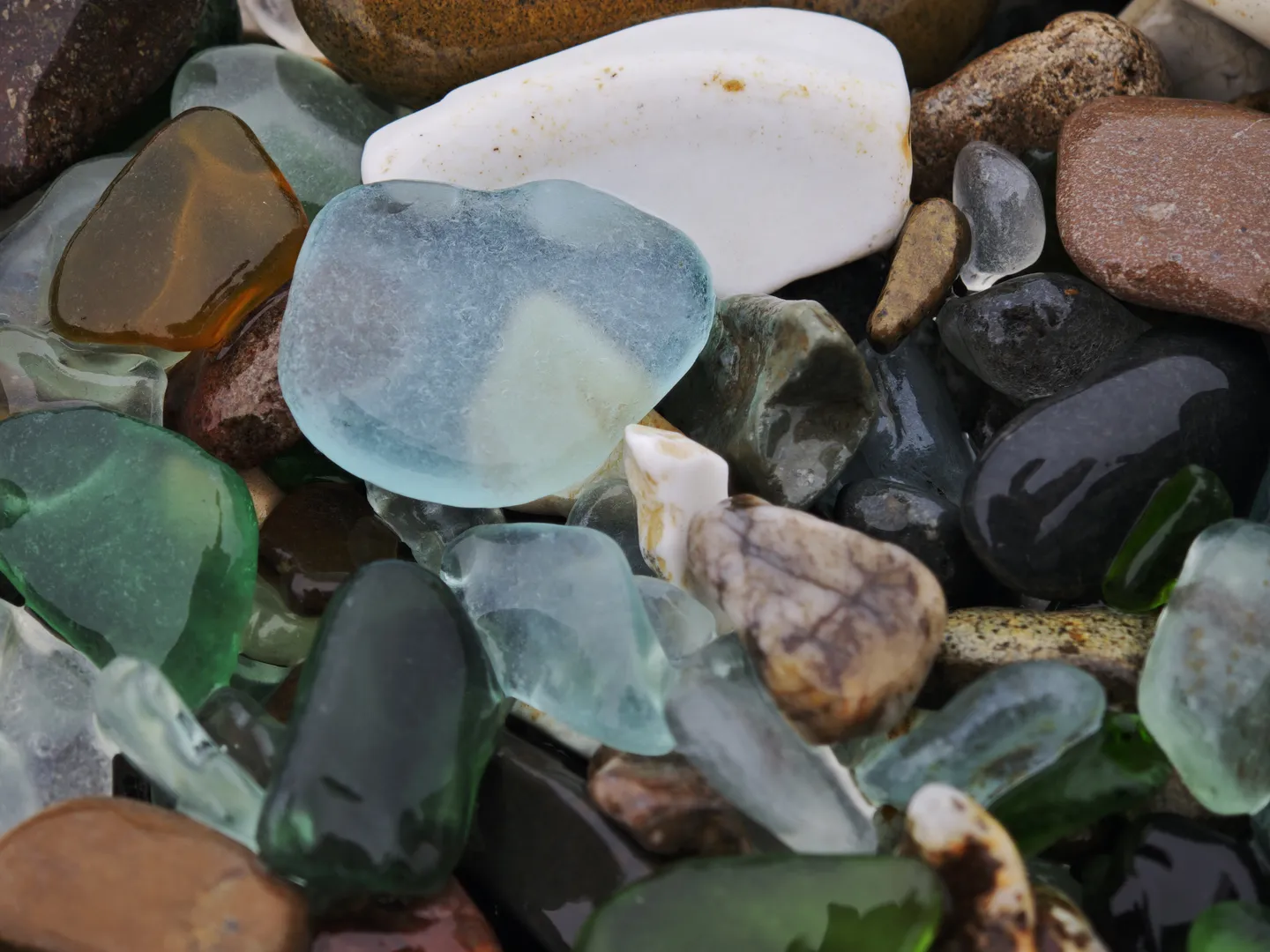 A pile of glass fragments and pebbles in rich colors, including blue, green, brown, and clear. The glass fragments are irregularly shaped, and the pebbles are mixed with them.