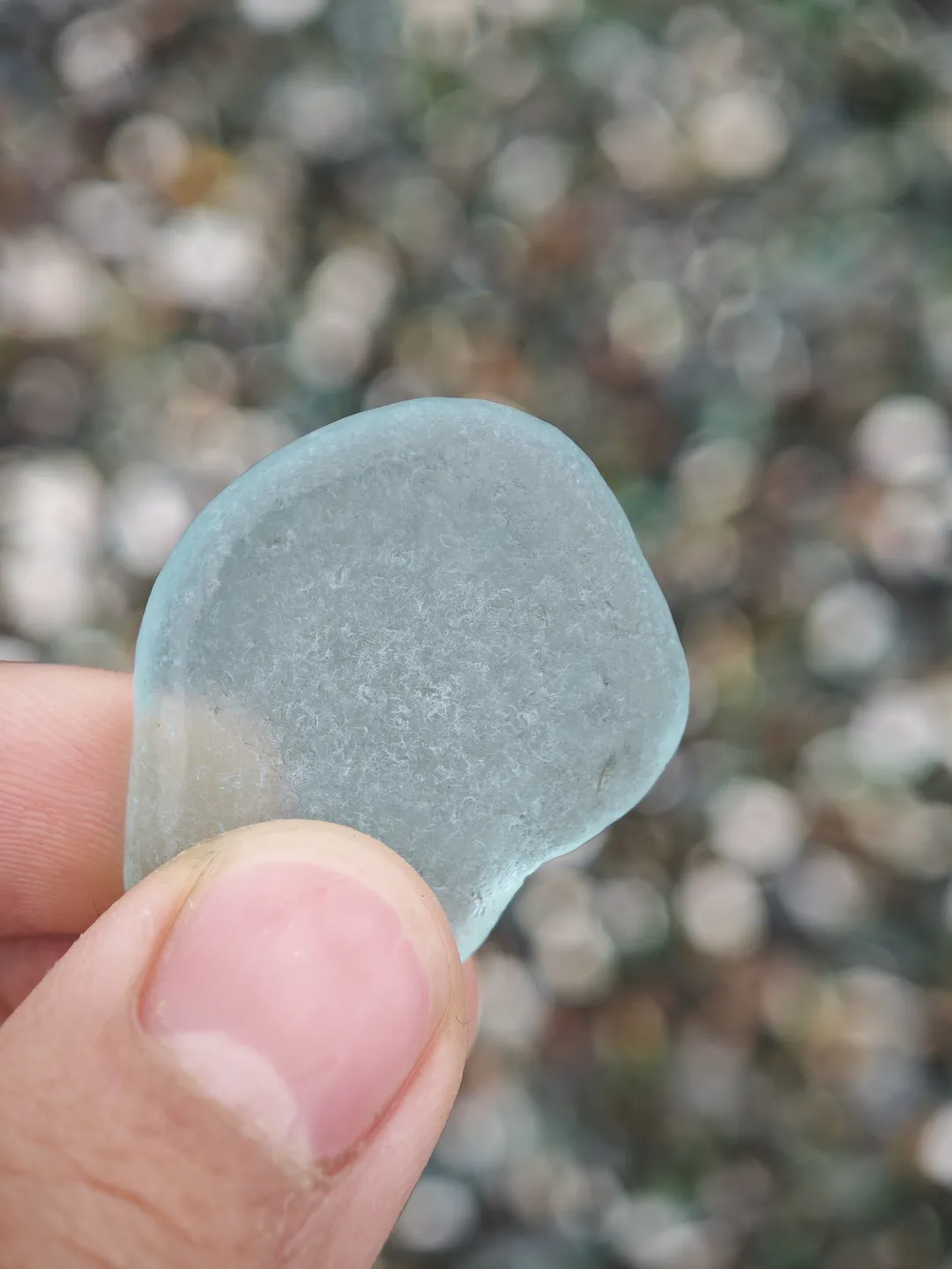 A hand holds a light blue glass fragment, with a blurred background. The glass is circular, smooth on the surface, and the fingers gently grip the edges.