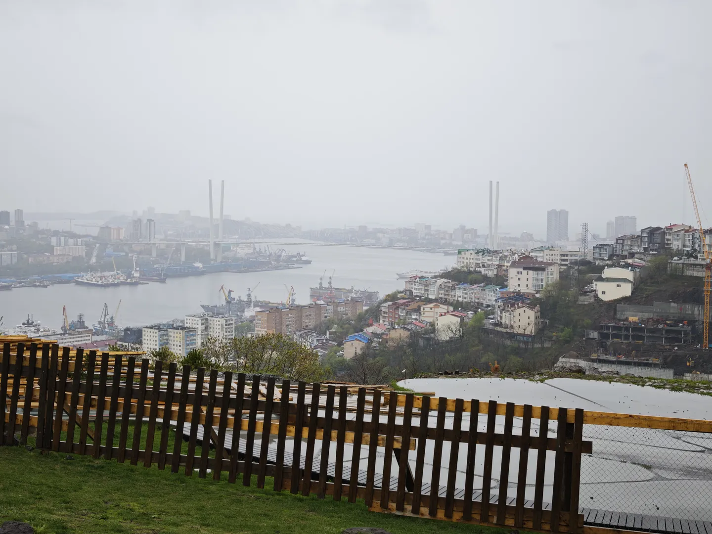 A cityscape viewed from above, with a wooden fence in the foreground. The background features dense buildings and a river. There is a bridge over the river, and both banks are filled with high-rise buildings and cranes. The sky is cloudy.