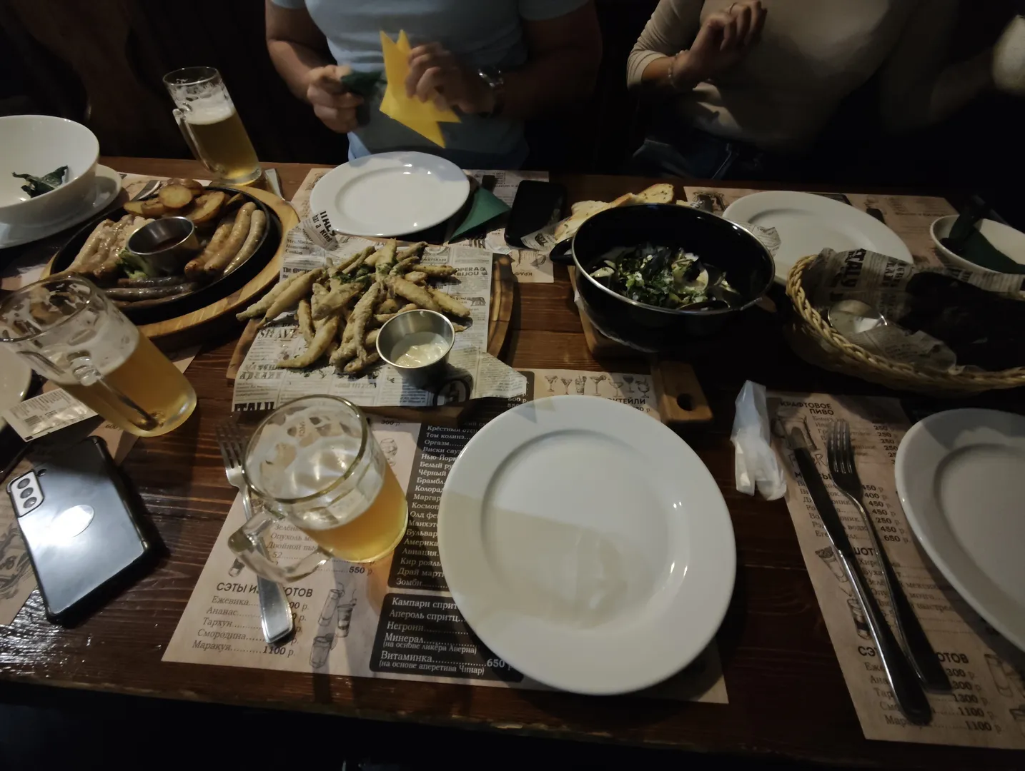 The dining table is set with various foods and drinks, including beer, grilled sausages, and salad. There is also a menu and a phone on the table, with several people sitting nearby. The atmosphere is dim, with soft lighting.