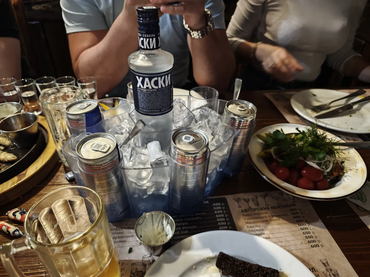 The dining table is set with various drinks and foods, with a bottle labeled “ХАСКИ” in the center. There are canned drinks, a bowl of salad, empty glasses, and ice cubes. Several people are seated at the table, with different hand gestures.
