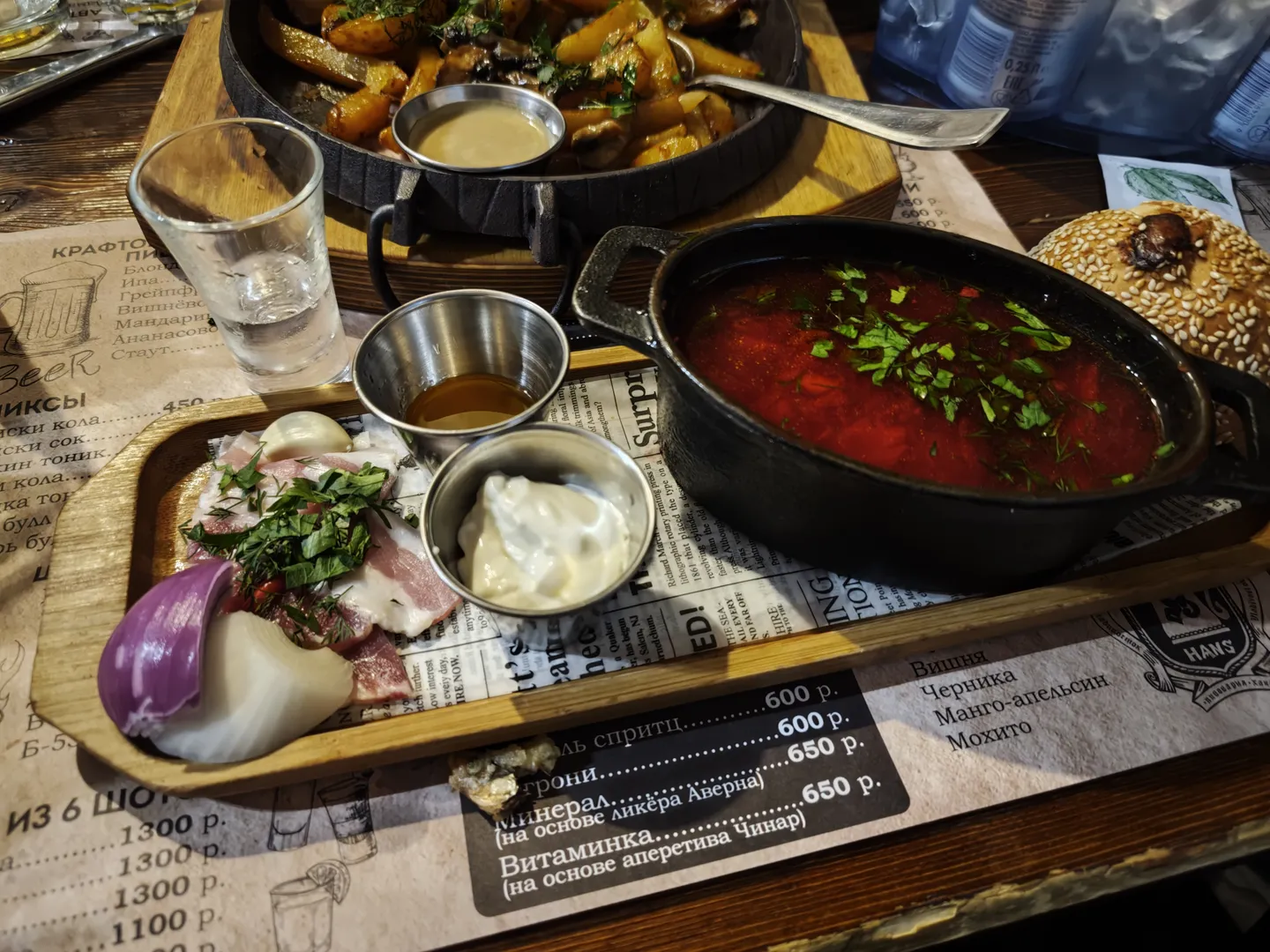 The dining table holds a pot of red soup, with sliced onions and seasonings on the side. The soup is served in a black pot, surrounded by small bowls of ingredients. A menu paper lists prices for various drinks.