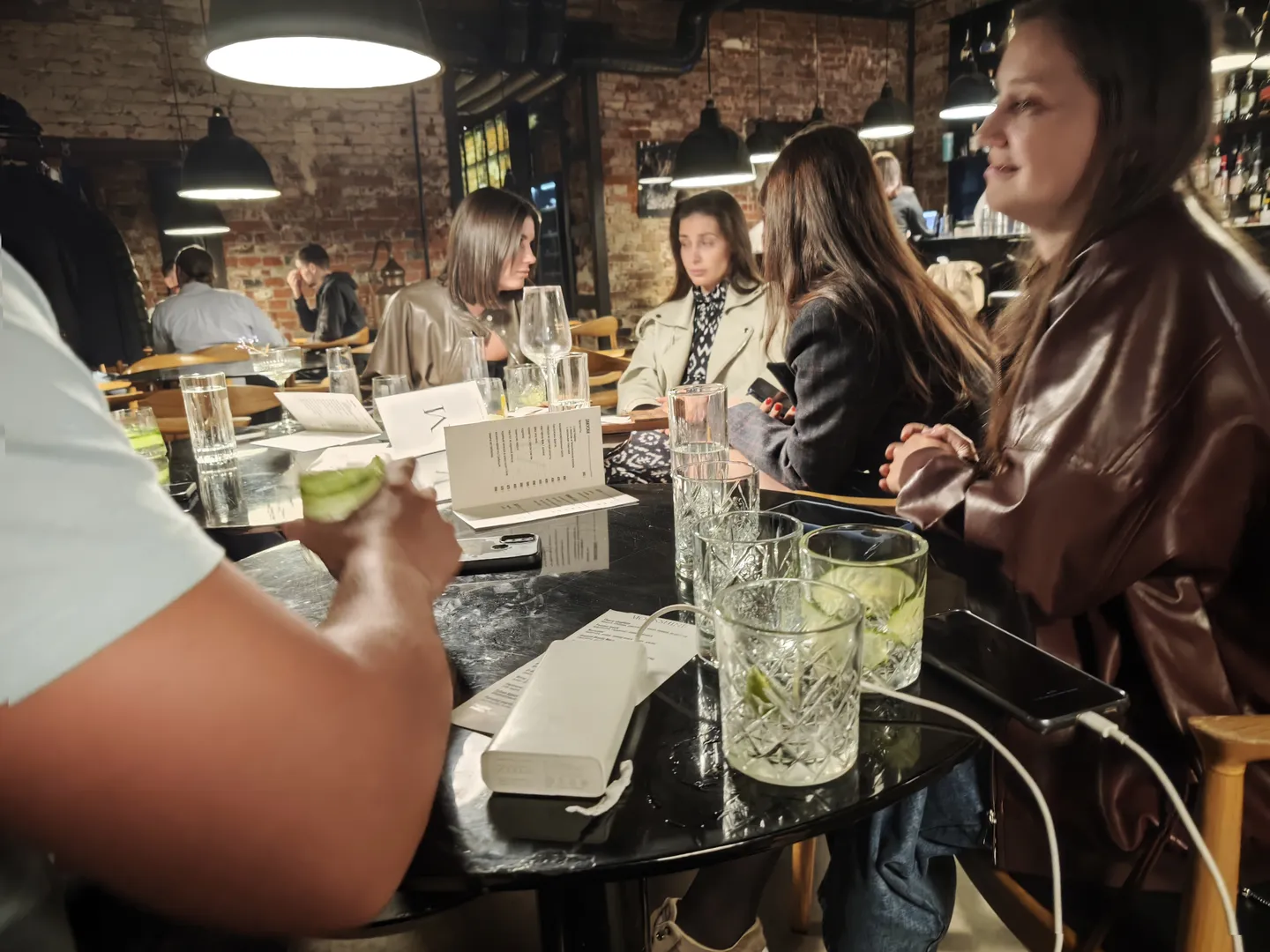 Several people are seated around a dining table in a restaurant. The table is set with transparent glass cups and a menu. A man is placing a slice of lemon into a cup, while several women sit across from him. The restaurant’s interior is modern, with warm lighting.
