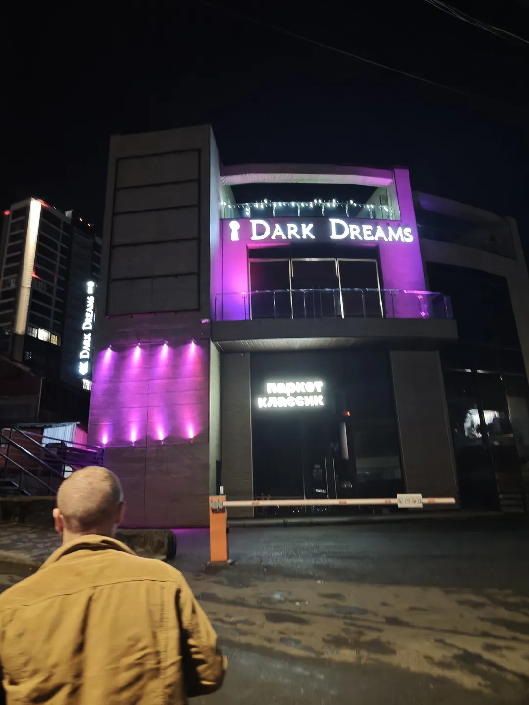 The exterior of the building at night, with the words “Dark Dreams” on the front and the “паркет классик” logo below. The exterior lights of the building are purple, and there are orange railings at the door. A man stands with his back to the camera.