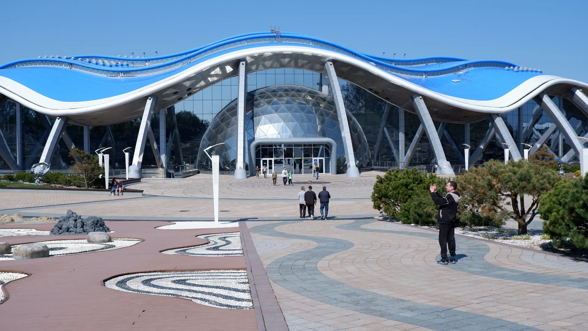 A modern building features a wavy blue roof and glass curtain wall, with several people walking on the plaza in front. At the entrance stands a large spherical structure surrounded by trees. The plaza ground is designed with curved patterns and stone ornamentation.