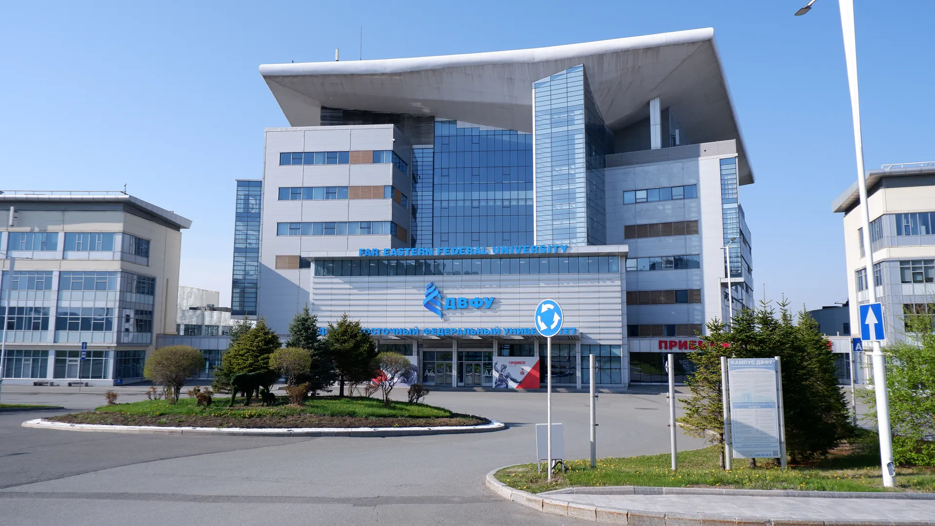 A tall building marked with the words “FAR EASTERN FEDERAL UNIVERSITY”, the exterior walls are made of glass and metal. In front, there is a green belt and bushes. To the left of the building, there is a lower building. To the right, there is a sign. The sky is clear, and other buildings are visible in the background.