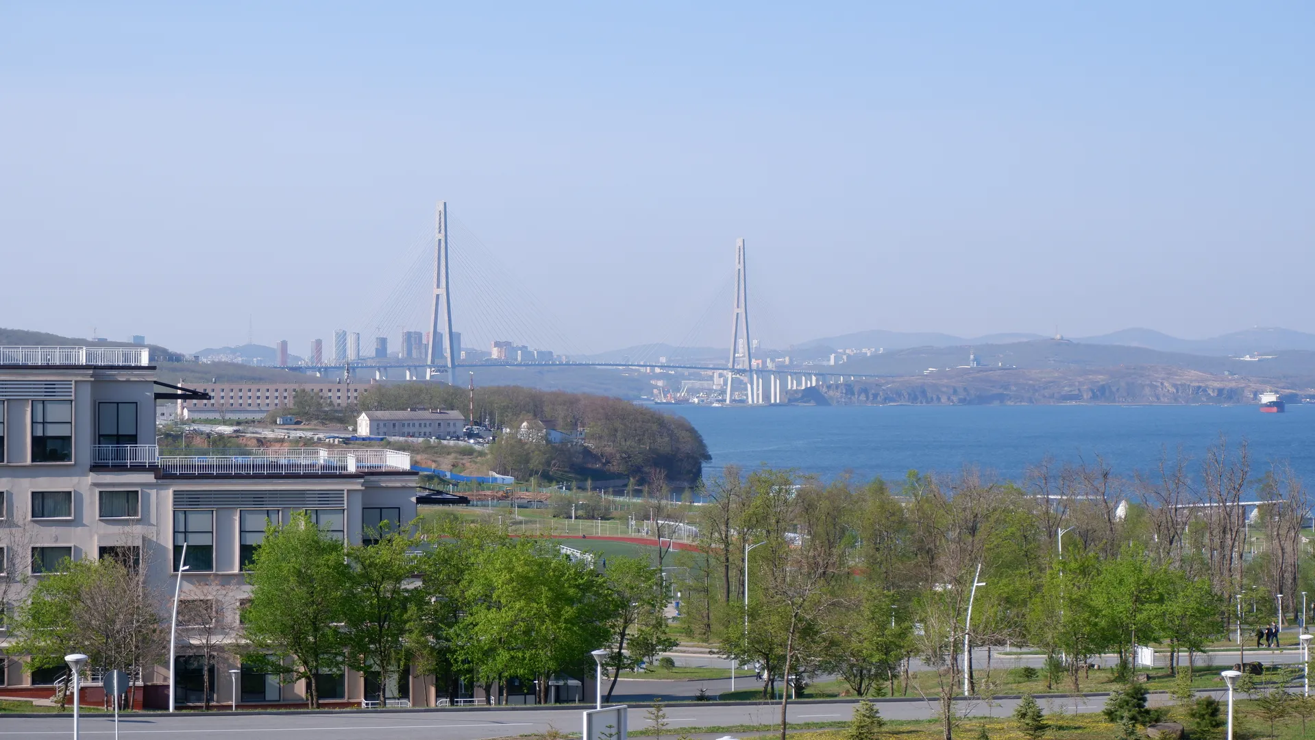 In the distance is a cross-sea bridge, connecting the urban landscapes on both sides. In the foreground, there are trees and some buildings, the water surface is calm. The sky is clear, and the view is expansive.