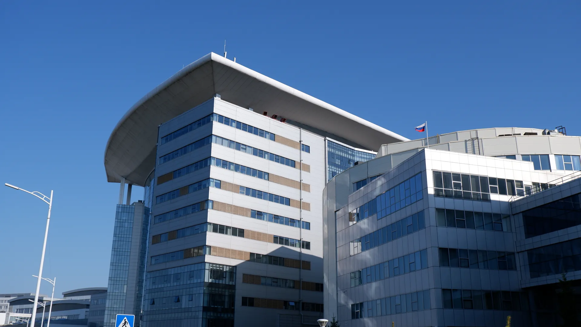 This is a modern high-rise building. The exterior walls are mainly composed of glass and metal. At the top of the building, there is a prominent curved structure with a flag fluttering on it. The sky is azure blue, without any clouds.
