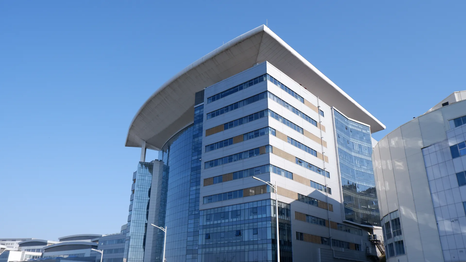 A modern high-rise building with large glass windows covering its facade. At the top of the structure, there is a distinctive curved element reflecting the sunlight. The sky is clear, and other buildings are visible in the background.