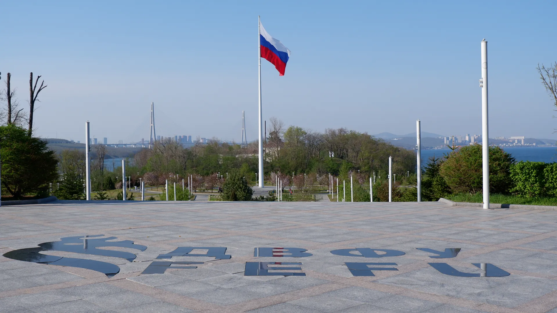 A square is paved with large stone bricks, in the center stands a tall flagpole on which flies the Russian flag. The ground of the square is engraved with the capital letters “ДВФУ”, surrounded by greenery and trees. In the distance, a cross-sea bridge and the city skyline can be seen.