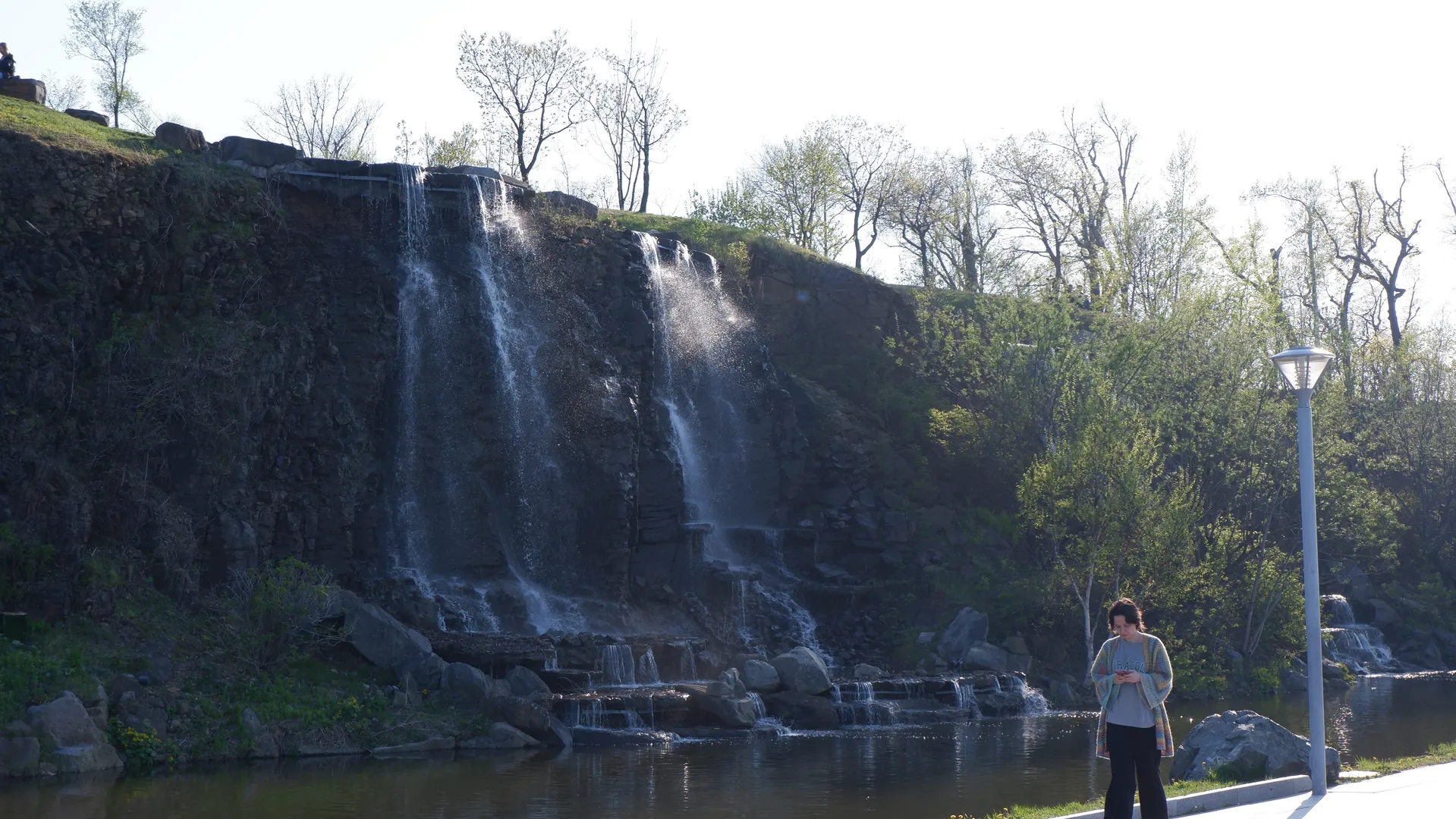 A waterfall cascades down from the cliff, separating into multiple streams. Below the waterfall lies a pond, beside which a person is standing and looking at his phone. Surrounding the area are dense trees and bushes.