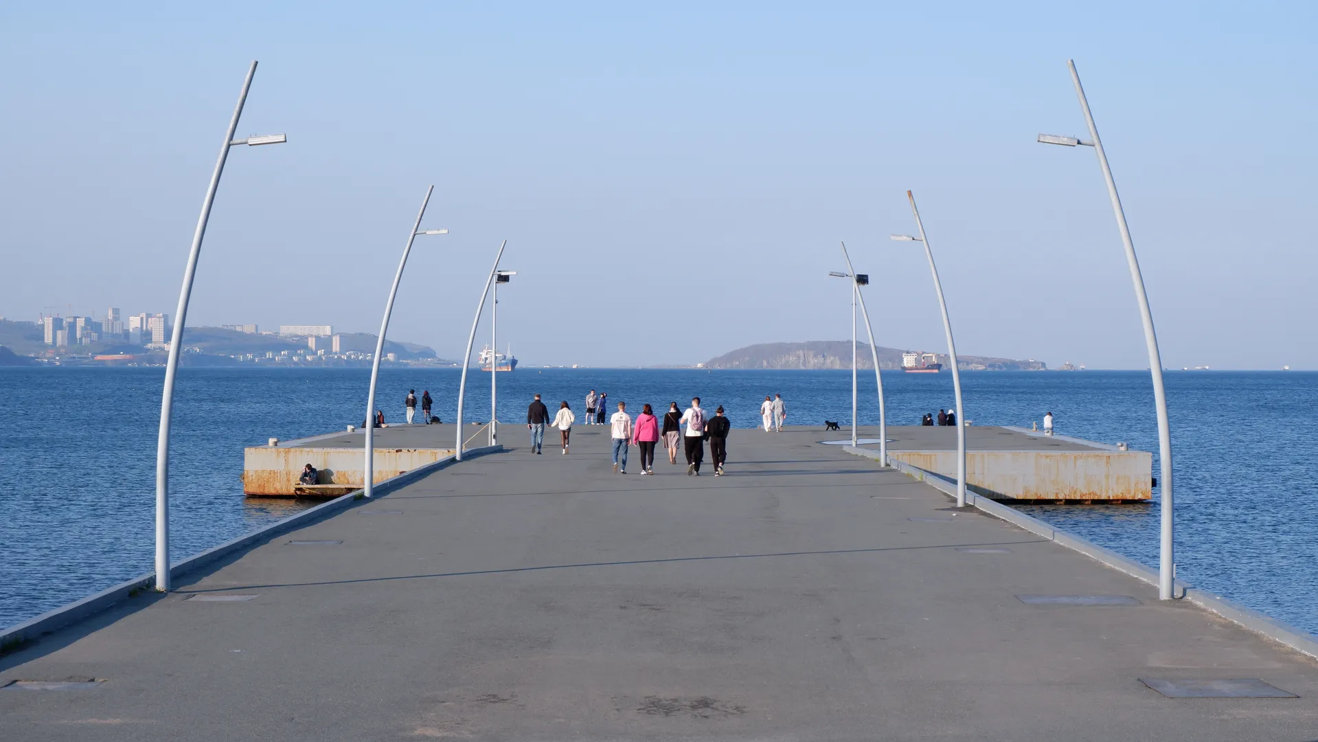 A straight coastal walkway extends into the sea, flanked by arched streetlights on both sides. Pedestrians are walking along the path, while several boats are anchored on the distant sea surface. The sky is clear, offering expansive views.