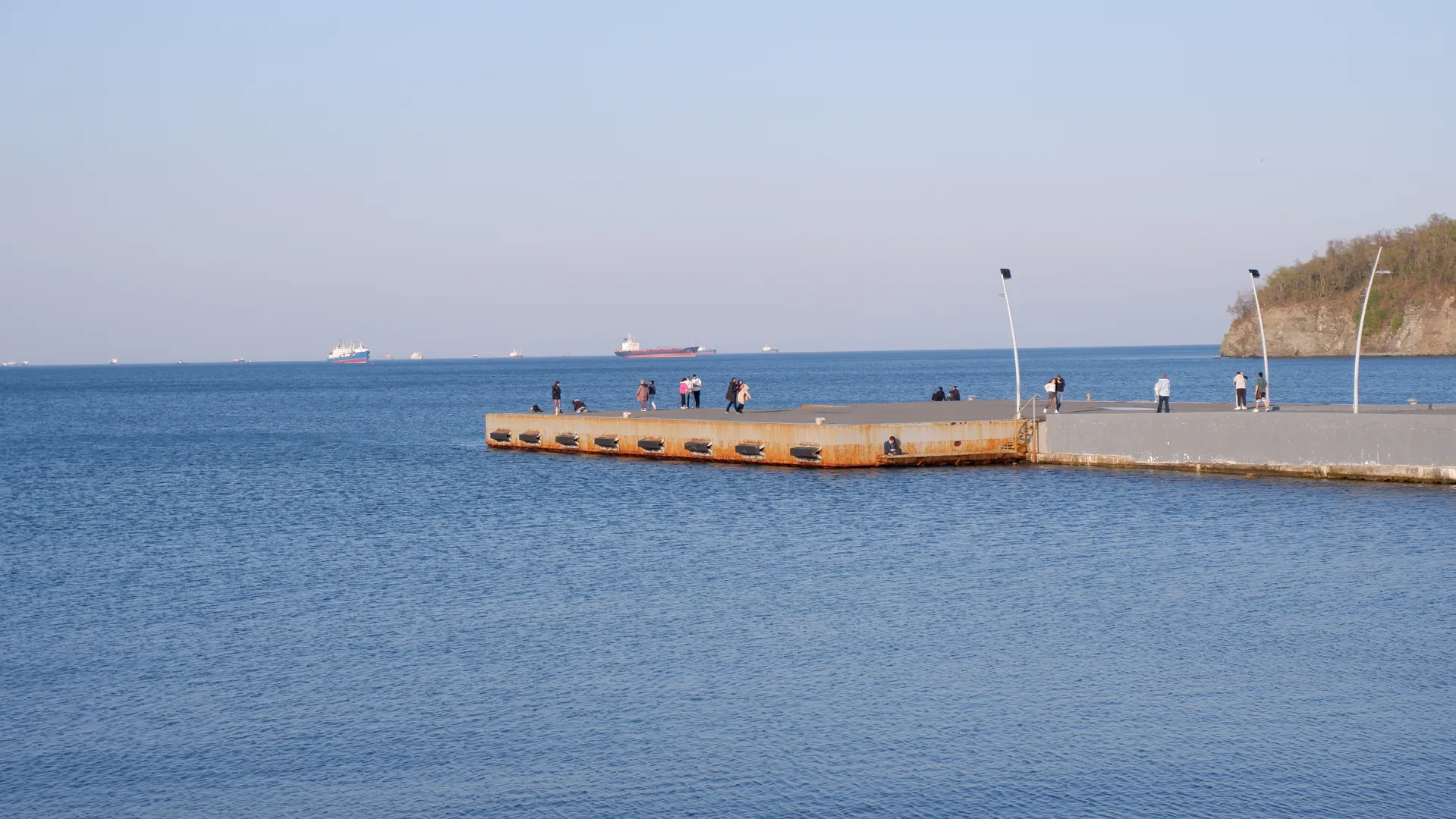 A vast expanse of water, with a platform extending into the sea at its center, where people are active. On the distant sea, several large ships are sailing. To the right is a small hill. The sky is clear, and the sea is calm.