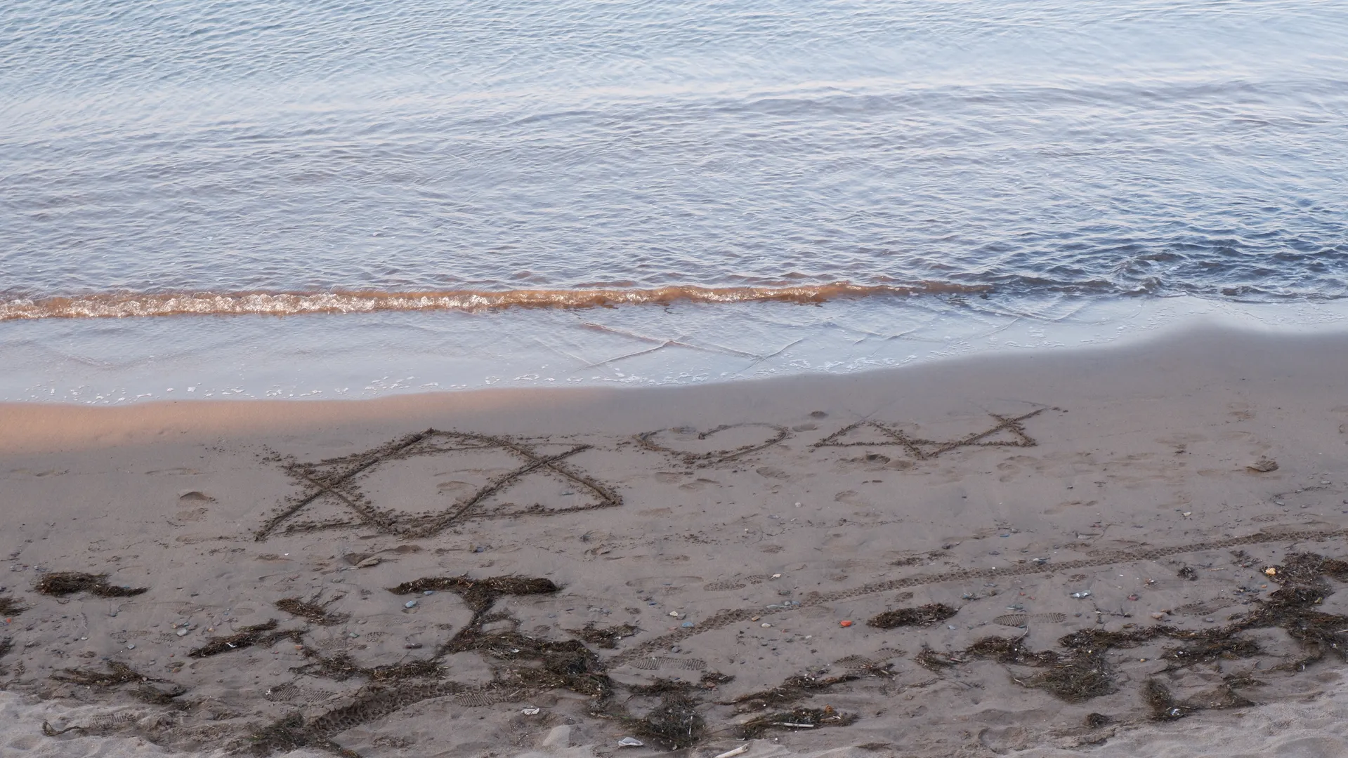 On the beach, two hexagrams and a heart symbol were drawn in the sand, with seawater gently lapping against the shore. Scattered seaweed and pebbles dotted the sandy shore. The calm surface of the distant waters reflected the light of the sky.