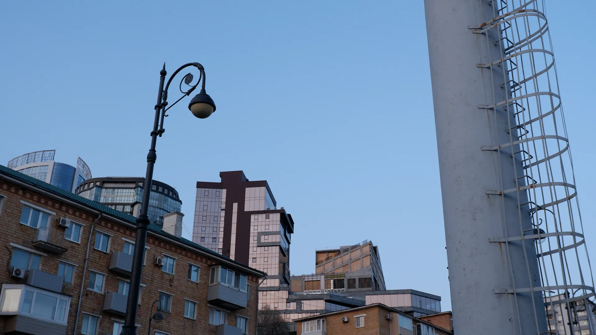 A vintage-style street lamp stands beside the city road, with multiple high-rise buildings in the background. The buildings feature modern designs with glass curtain walls on their facades. The sky is clear, presenting a pale blue hue.