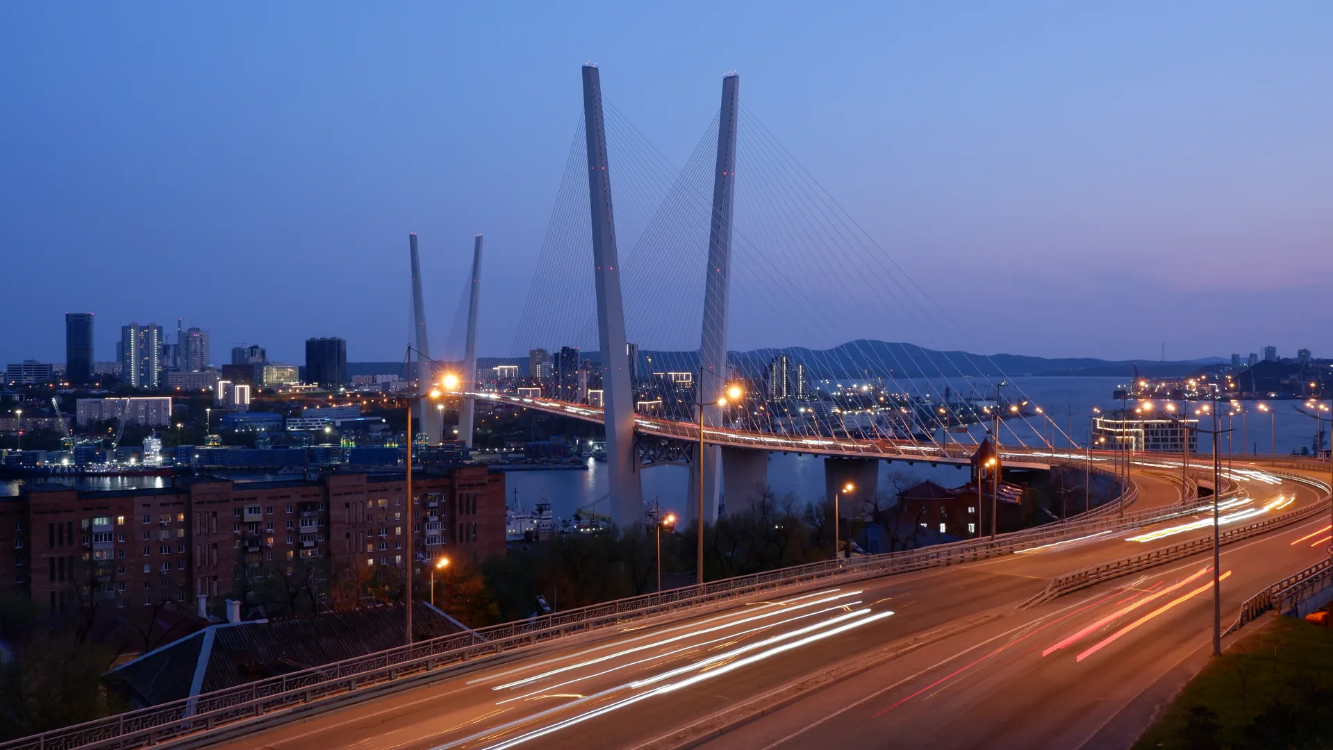 The city bridge glows brilliantly at night, with light trails left by passing vehicles on its surface. The structure features a cable-stayed design, with towering pillars. In the distance, the city lights and the outline of mountains are visible.