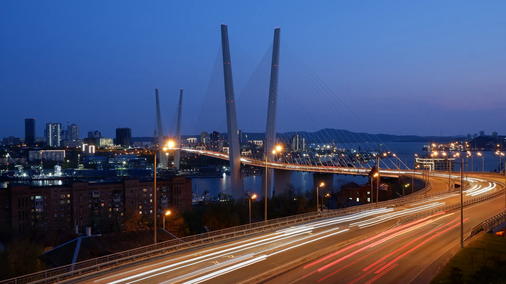 The city bridge at night is brilliantly illuminated, with light trails left by vehicles traversing its span. The bridge features a cable-stayed design, with towering pylons rising above. In the distance, the lights of the city and the silhouettes of mountains are visible against the horizon.