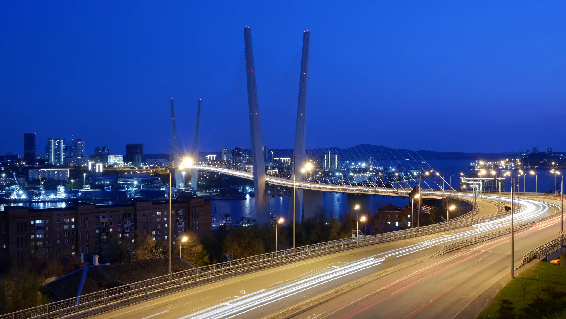 The city bridge at night is brilliantly lit. On the bridge, there are light trails left by moving vehicles. The bridge structure is of a cable-stayed design, with towering pylons. In the distance, the city lights and mountain silhouettes are visible.