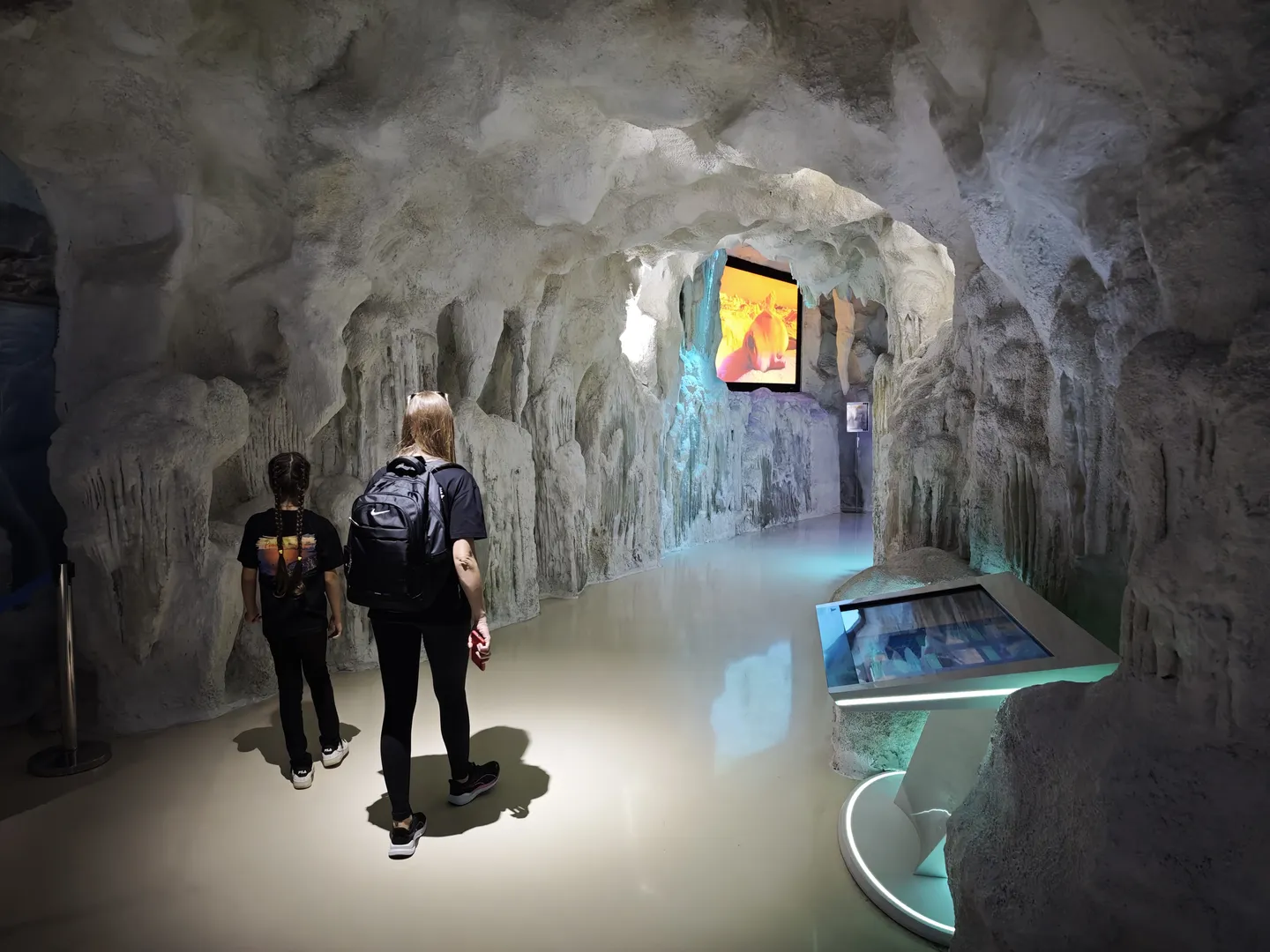 In a simulated cave interior scene, two people are visiting. The cave walls have stalactites and stalagmites, and there is a display screen in front playing related content. The ground is smooth, and the ambient lighting is soft.