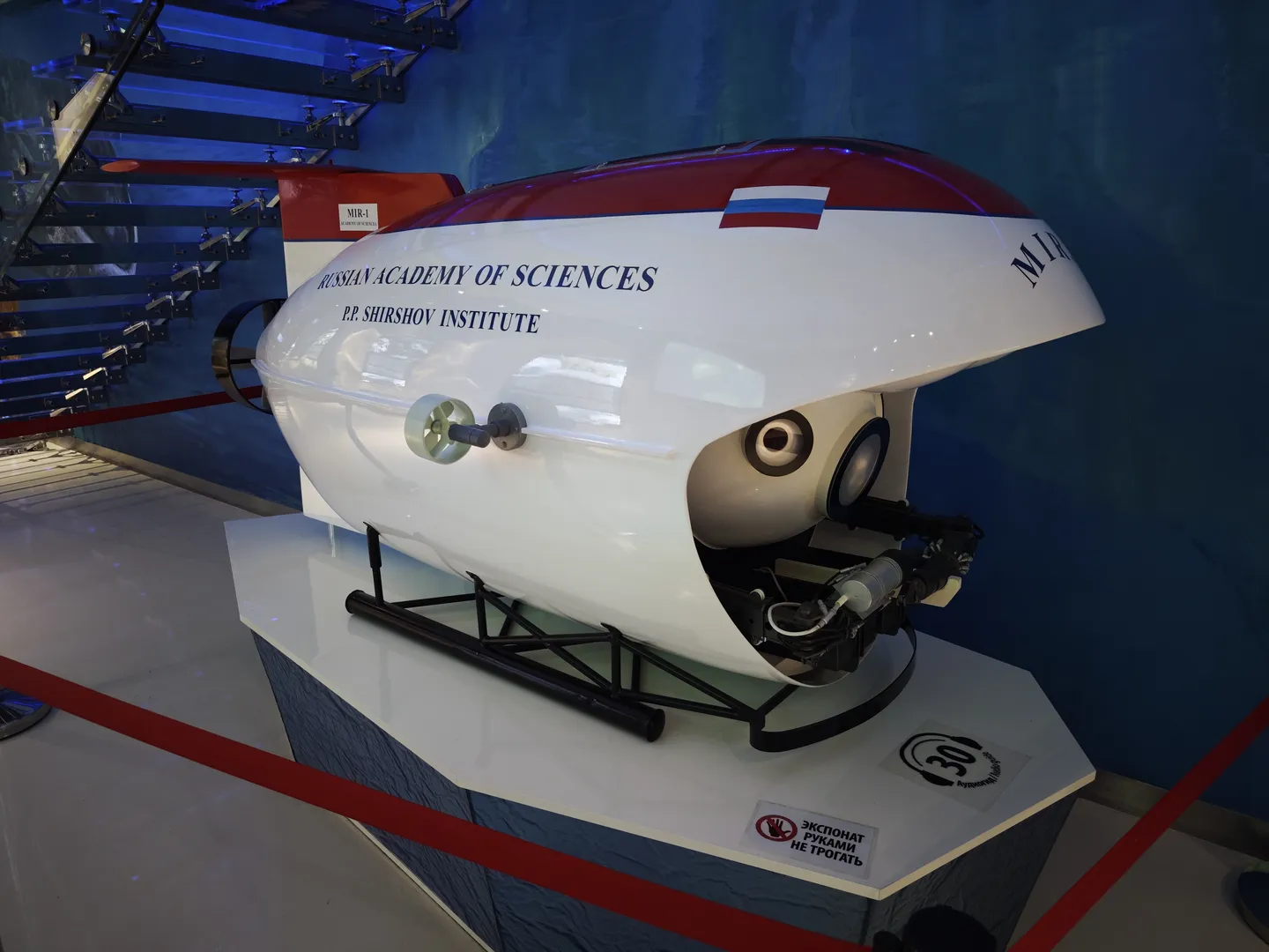 A model of a submersible labeled “RUSSIAN ACADEMY OF SCIENCES P.P. SHIRSHOV INSTITUTE” is displayed on the exhibition stand. Featuring a streamlined design, the model has a white main body adorned with red and blue stripes. A “Do Not Touch” sign is placed beside it.