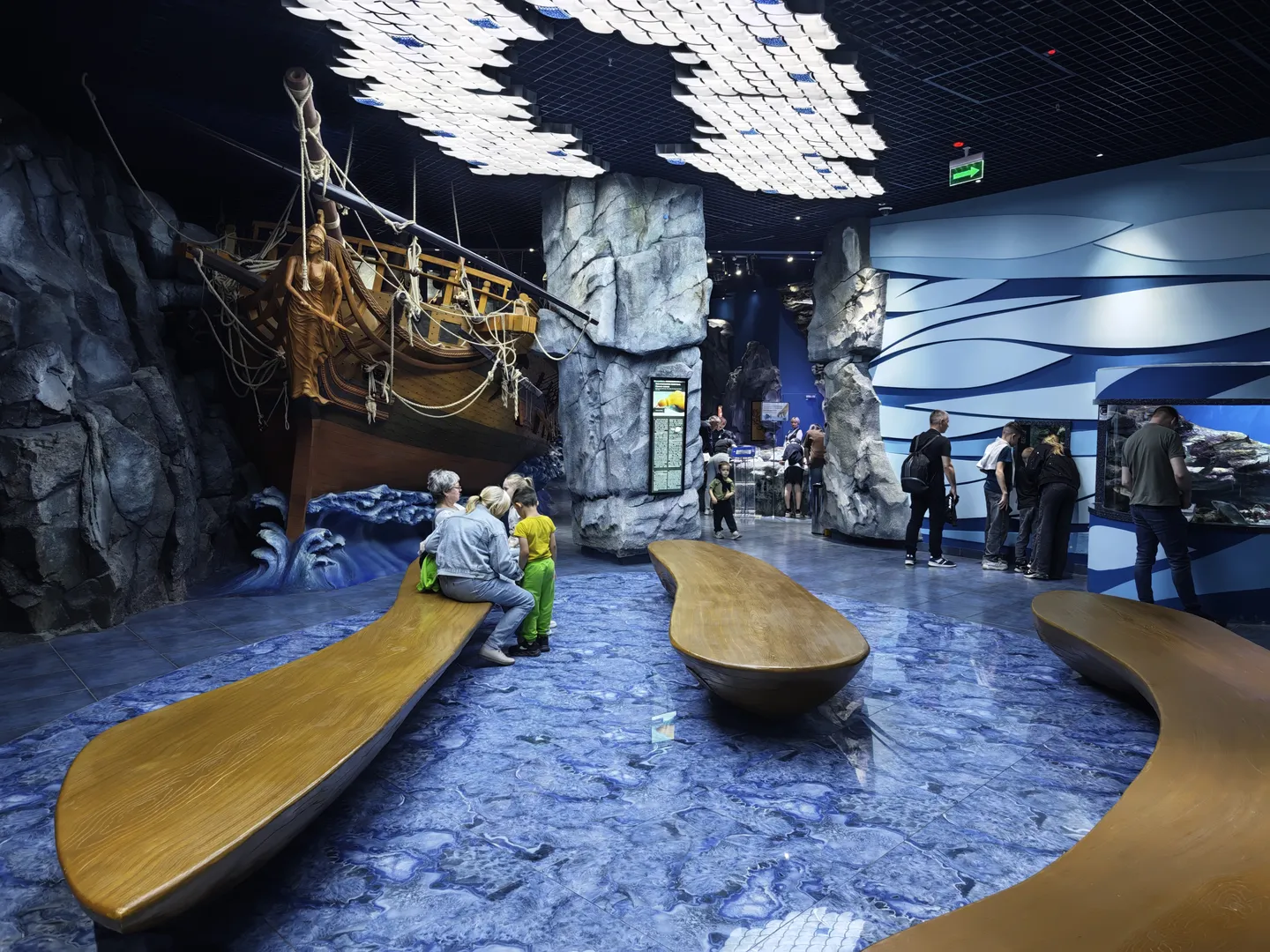 Inside the exhibition hall, there are wooden benches. The floor is decorated with blue wave patterns. On the left side, there is a large model of a ship, with a background of simulated rocks and waves. Several visitors are sitting on the benches or moving around inside the exhibition hall.