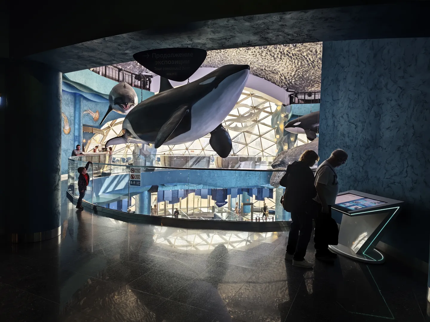 Several large whale models are hanging in the indoor exhibition space. There is a sign on the ceiling that says “Продолжение экспозиции Tour continued here”. The floor is smooth and reflective. Several visitors are viewing the exhibits. In the background, there are stairs and more exhibition content.