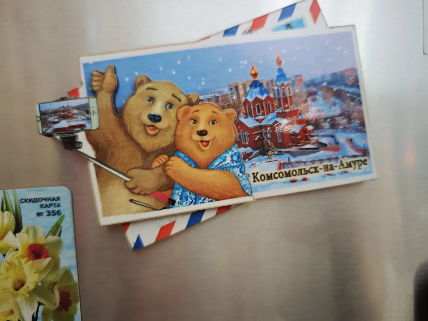 On a postcard, two cartoon bears are taking a selfie with a red church and snowy scenery in the background. The bottom right corner of the postcard reads “Комсомольск-на-Амуре”. Next to it is a card with a yellow flower pattern.