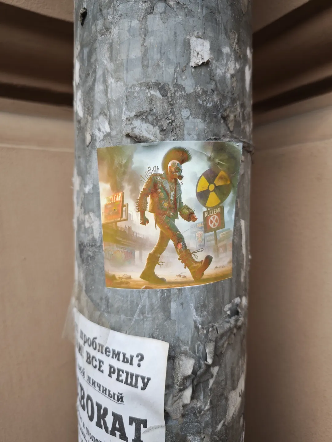 A poster is affixed to the pillar, depicting a figure in colorful attire with a “NUCLEAR” logo and radiation warning symbols in the background. Beneath the poster, there is a partially torn piece of paper with Russian text.