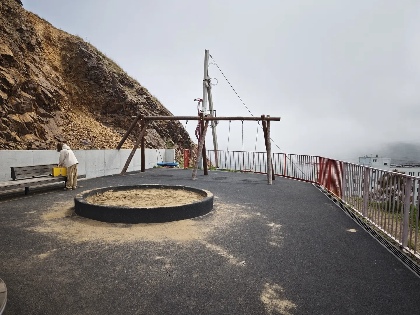 An empty playground with asphalt ground, featuring a sandpit and a swing set in the center. On the left, a person is bending over to place a yellow box, with a steep mountain wall in the background. In the distance, there are buildings and fences.