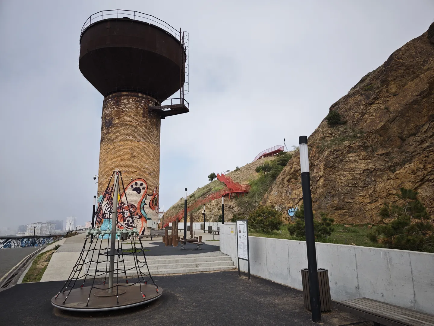 A large water tower stands at the center of the scene, with colorful graffiti on its surface. In front of the water tower is a children’s play facility, and to the right is a steep hillside with red stairs. In the foreground, there are benches and trash cans.