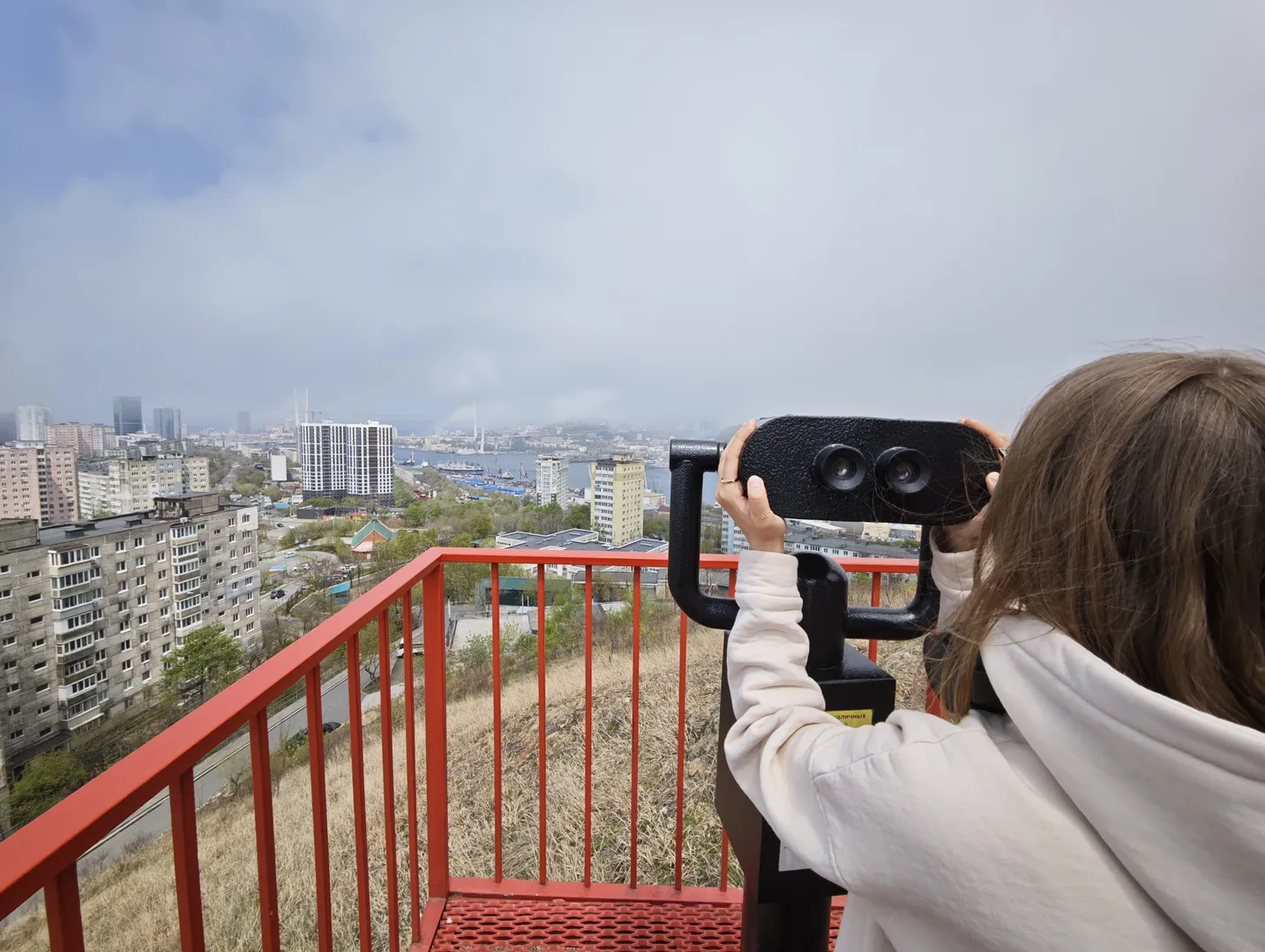 A person stands on an observation deck, observing the distant cityscape through binoculars. The observation deck has red guardrails, with dense buildings and green trees ahead. The sky is gloomy, with thick layers of clouds.