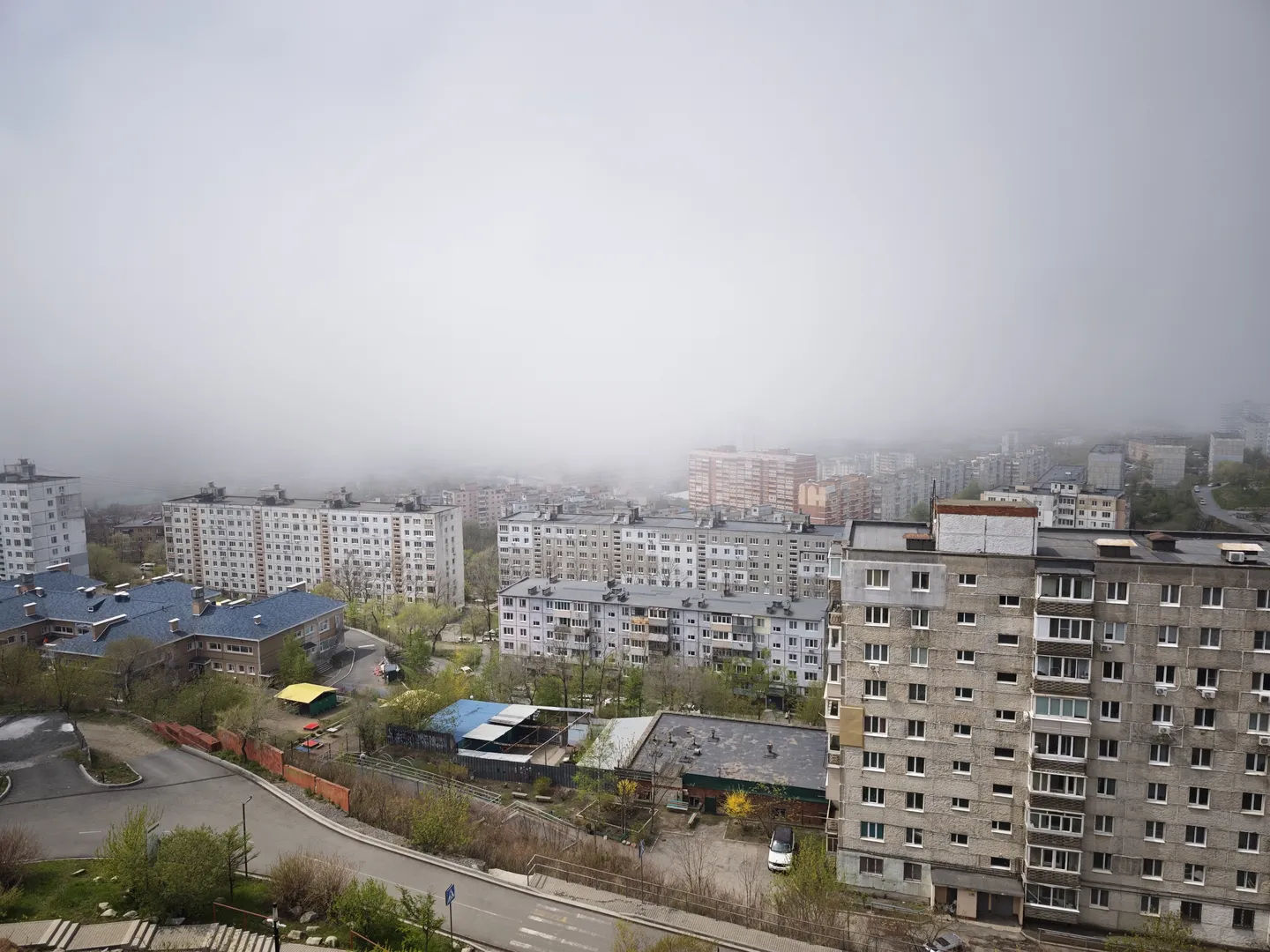 A residential area with neatly arranged multi-story buildings featuring diverse exterior colors. Roads and green belts run between the buildings, with vehicles moving along the roads. The sky is covered in thick fog, resulting in low visibility.