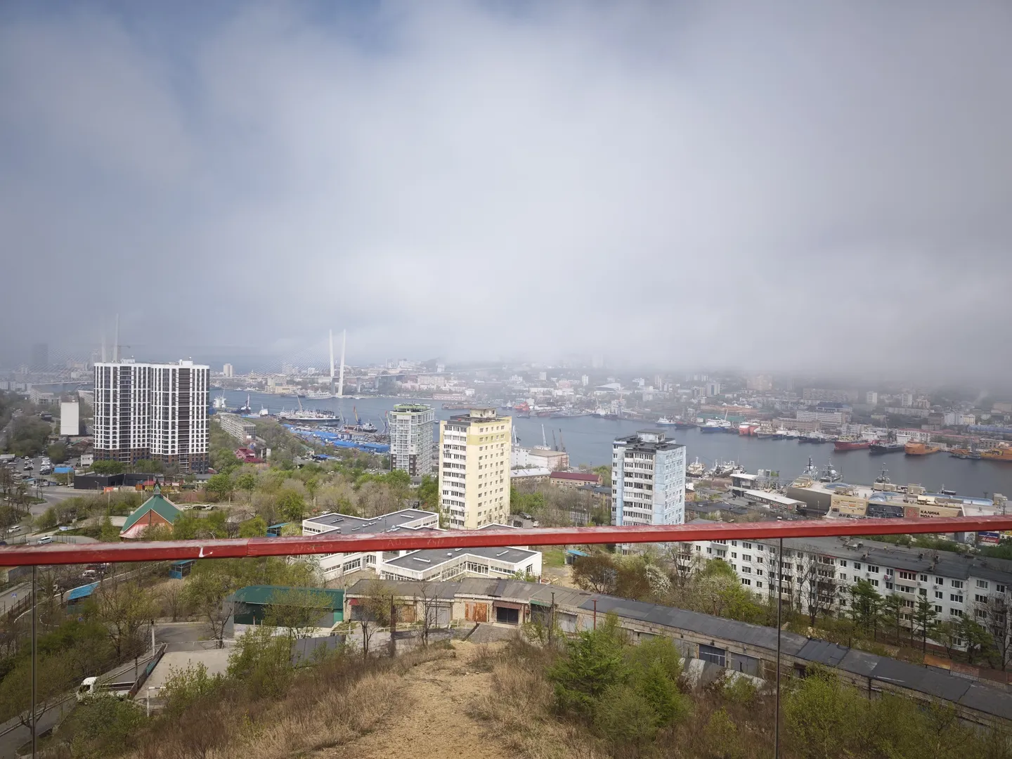 The panoramic view of the city is seen from a high vantage point, with low-rise buildings and greenery in the foreground, high-rise buildings and a river in the middle ground, and bridges and boats on the river. The sky is overcast, shrouded in clouds and mist, partially obstructing the view. There are red guardrails in the foreground.
