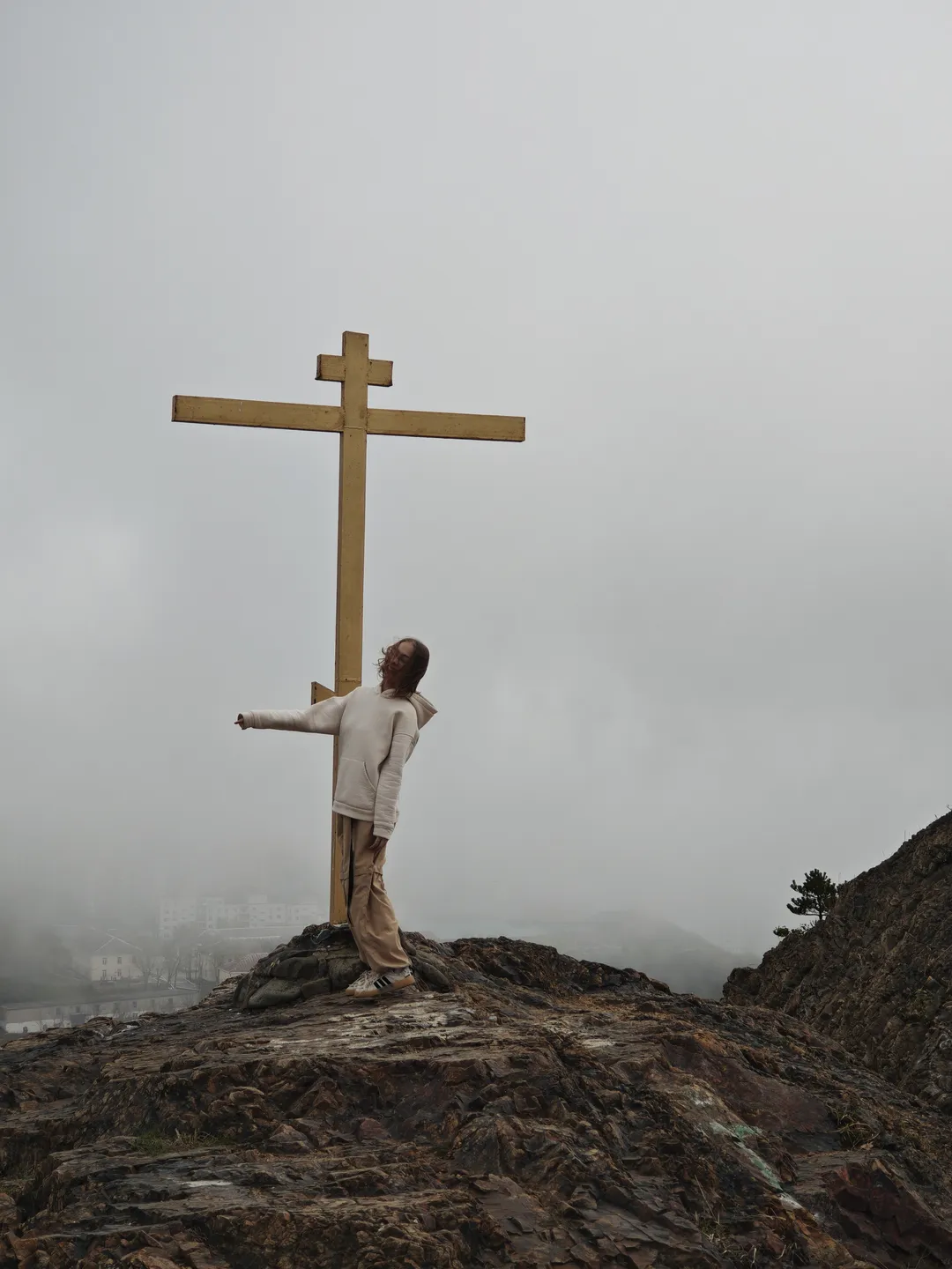 A person stands on a rock with a large cross beside them, against a backdrop of a hazy sky and distant buildings. The individual is dressed in light-colored clothing, raising their right hand to point to one side. The surface of the rock is rough, with sparse vegetation around.