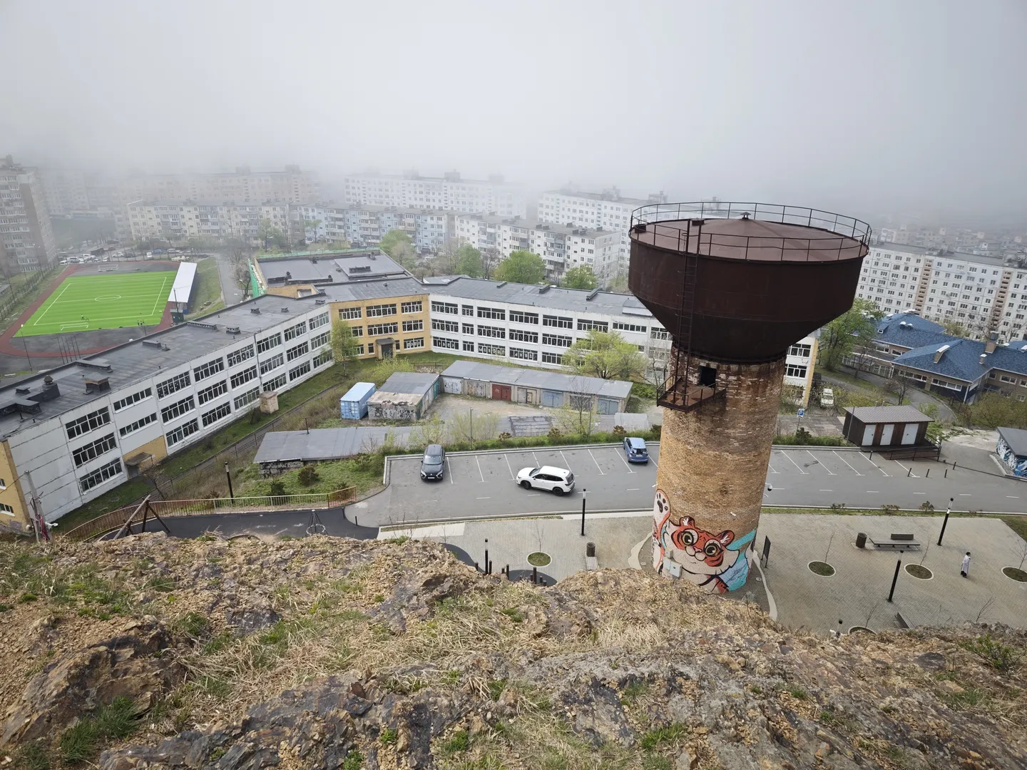 A tall water tower stands in the foreground, with colorful graffiti at its base, and multi-story residential buildings along with a football field in the background. Next to the water tower is a parking lot with several cars, and distant buildings are faintly visible through the mist. The foreground features an expanse of bare rocky ground.