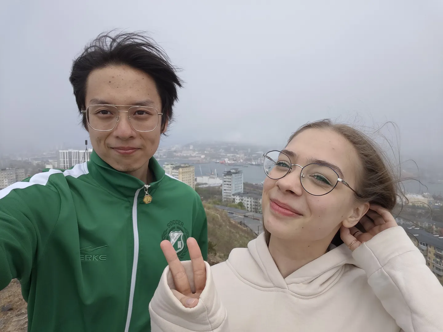Two people are taking a selfie from a high vantage point, with a cityscape and harbor in the background. The person on the left is wearing a green jacket, while the one on the right has a white hoodie, both smiling.