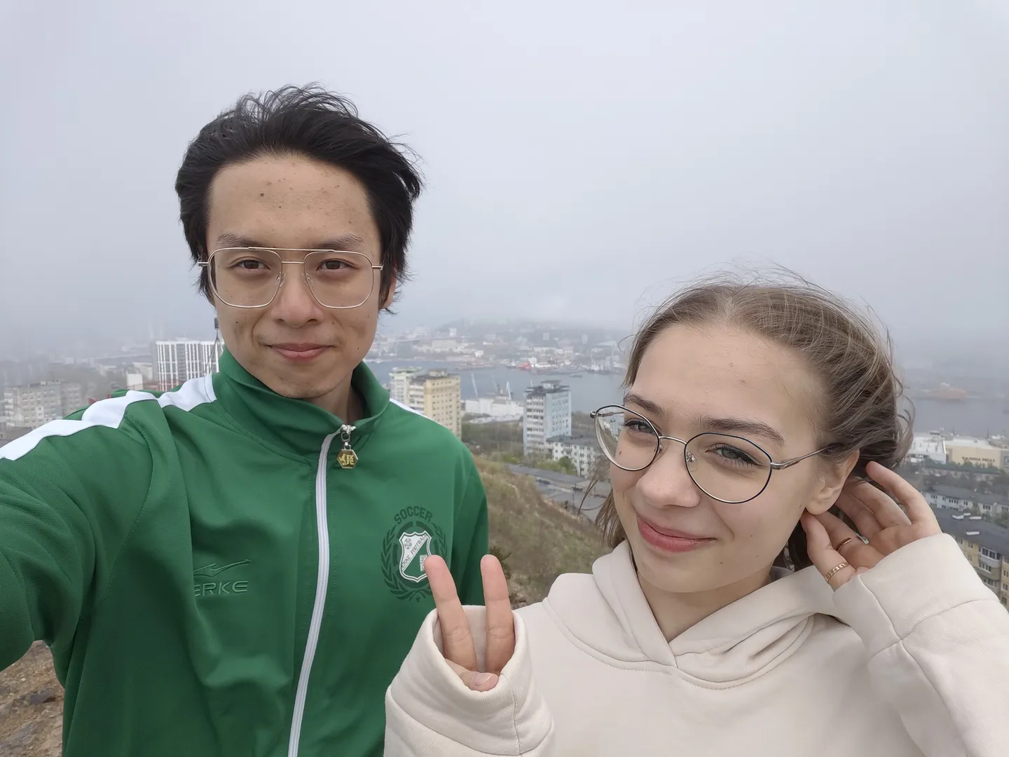 Two people are taking a selfie from a high vantage point, with a cityscape and harbor in the background. The person on the left is wearing a green jacket, while the one on the right has a white hoodie, both smiling.