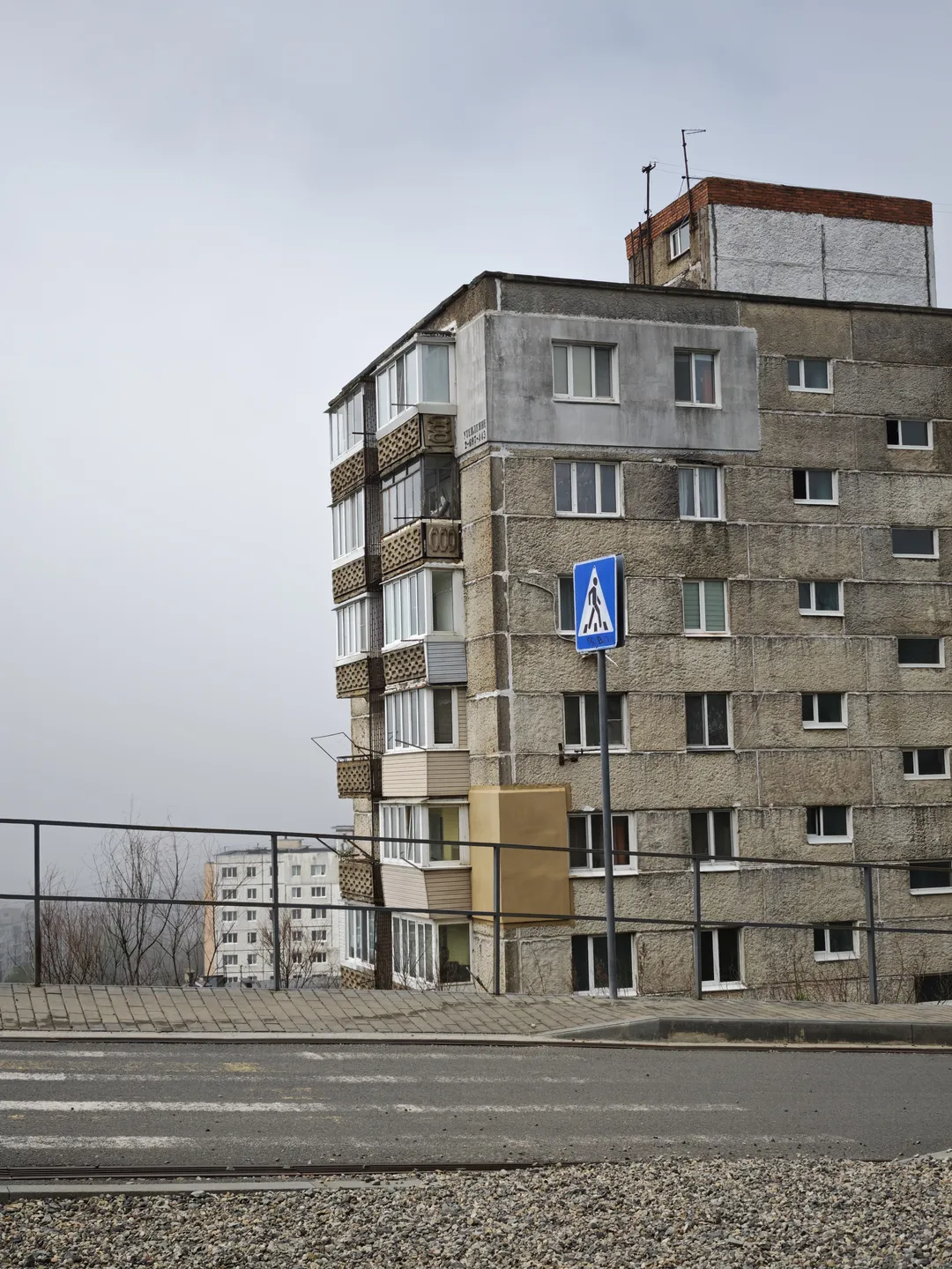 A multi-story building with gray concrete exterior walls, some balconies featuring decorative railings. There is a road in front of the building, with a pedestrian crossing sign on the roadside.