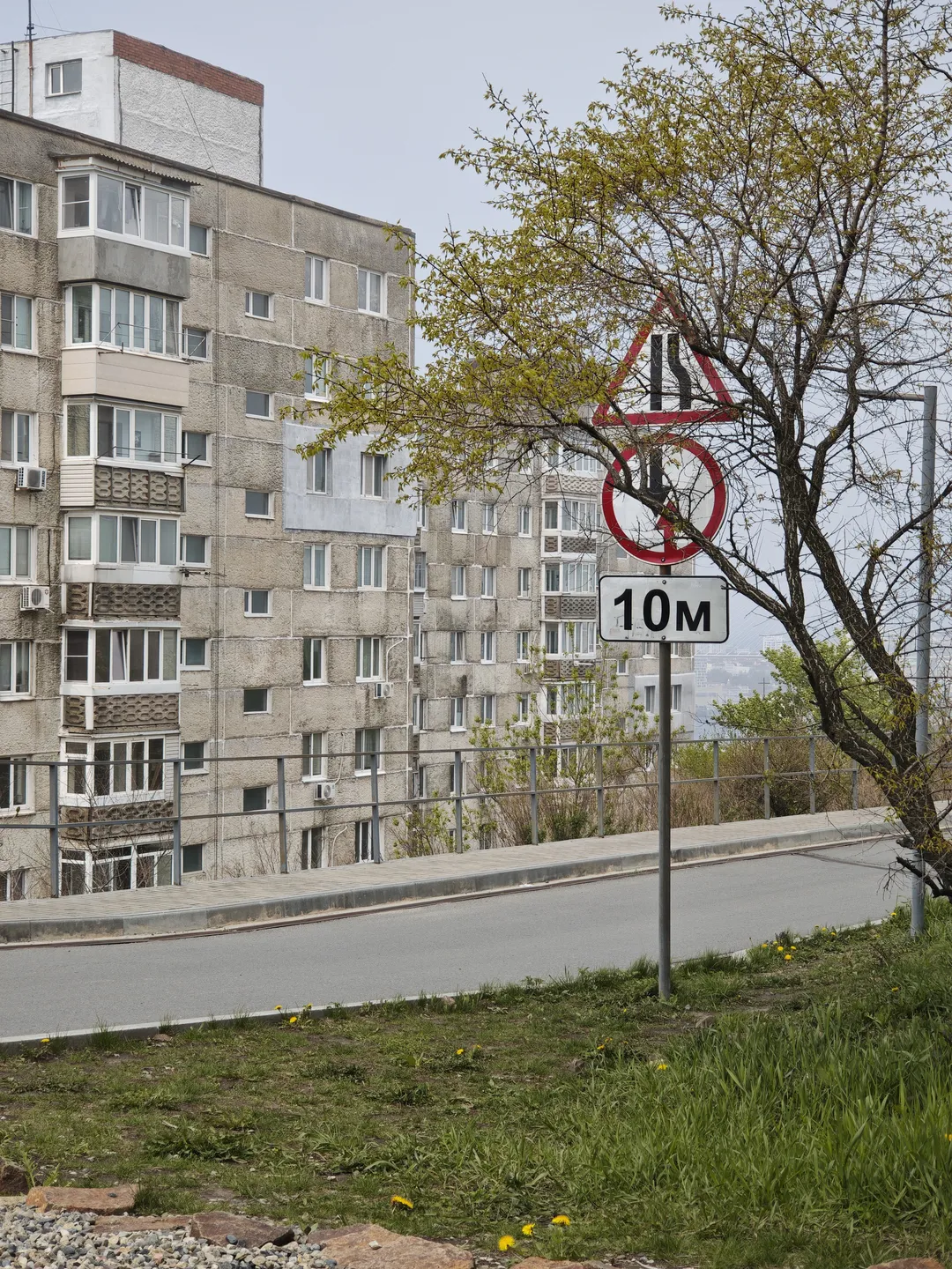 A multi-story building with gray concrete exterior walls, some balconies featuring decorative railings. There is a road in front of the building, with a traffic sign by the roadside indicating “No Honking” and a height limit of ten meters.