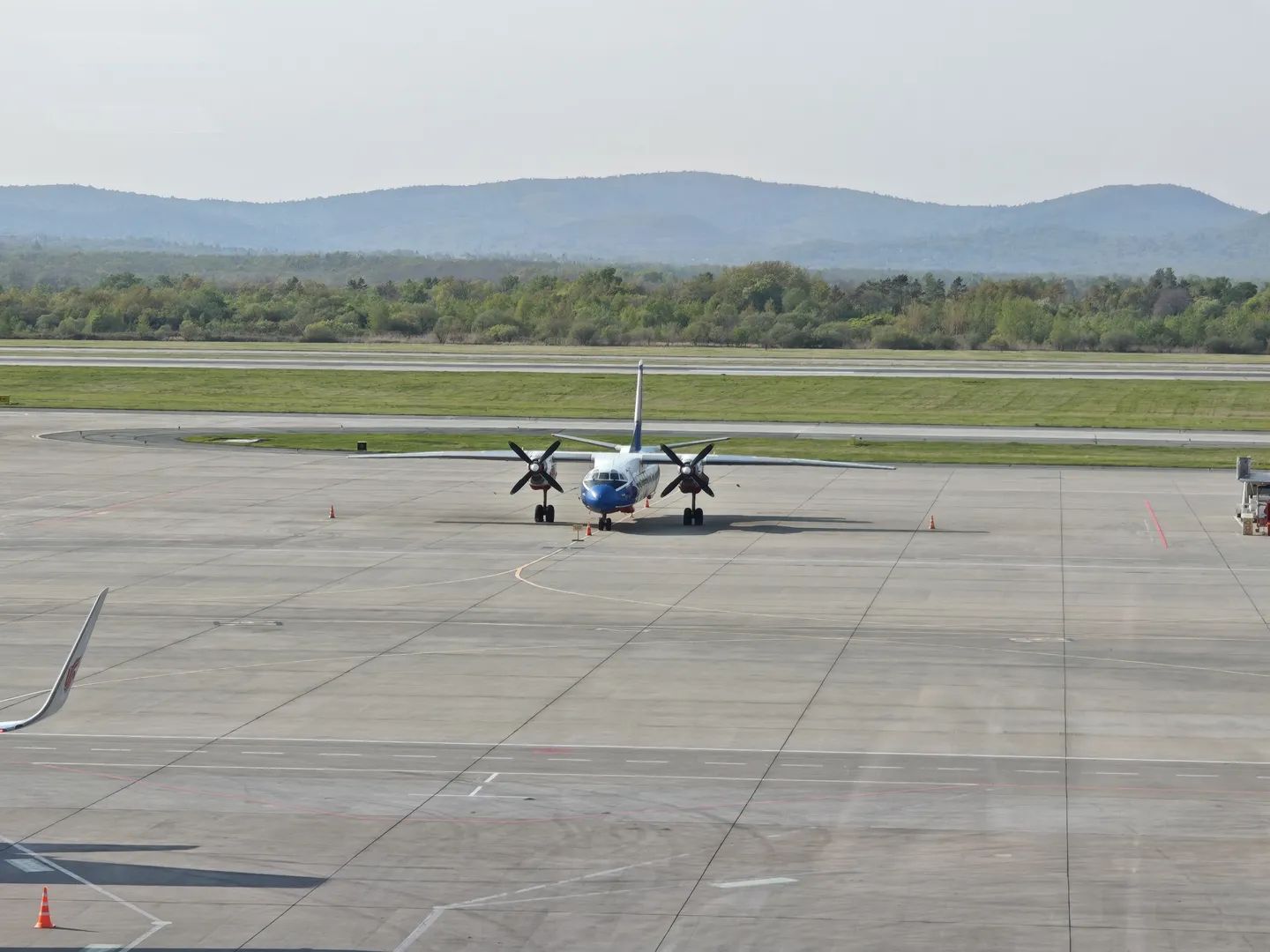 On the airport tarmac, a twin-engine propeller aircraft is parked, with a wide runway ahead. In the distance, there are dense trees and rolling mountains. The tarmac is marked with clear lines.