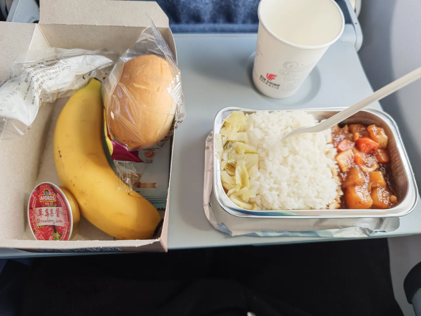The airplane meal tray includes a serving of rice, stew, and kimchi, alongside a banana, a bread roll, and a small jar of strawberry jam. There is also a paper cup printed with the “Air China” logo.