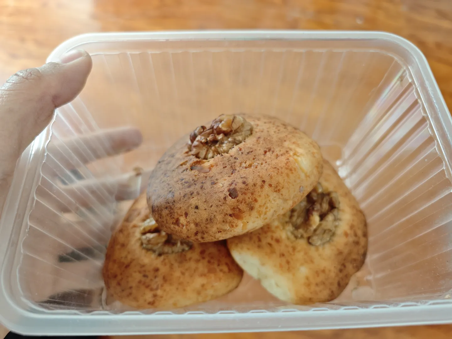 Inside the transparent plastic box are three round pastries, sprinkled with crushed nuts on the surface. The pastries are golden in color and neatly shaped. A hand is holding the plastic box.