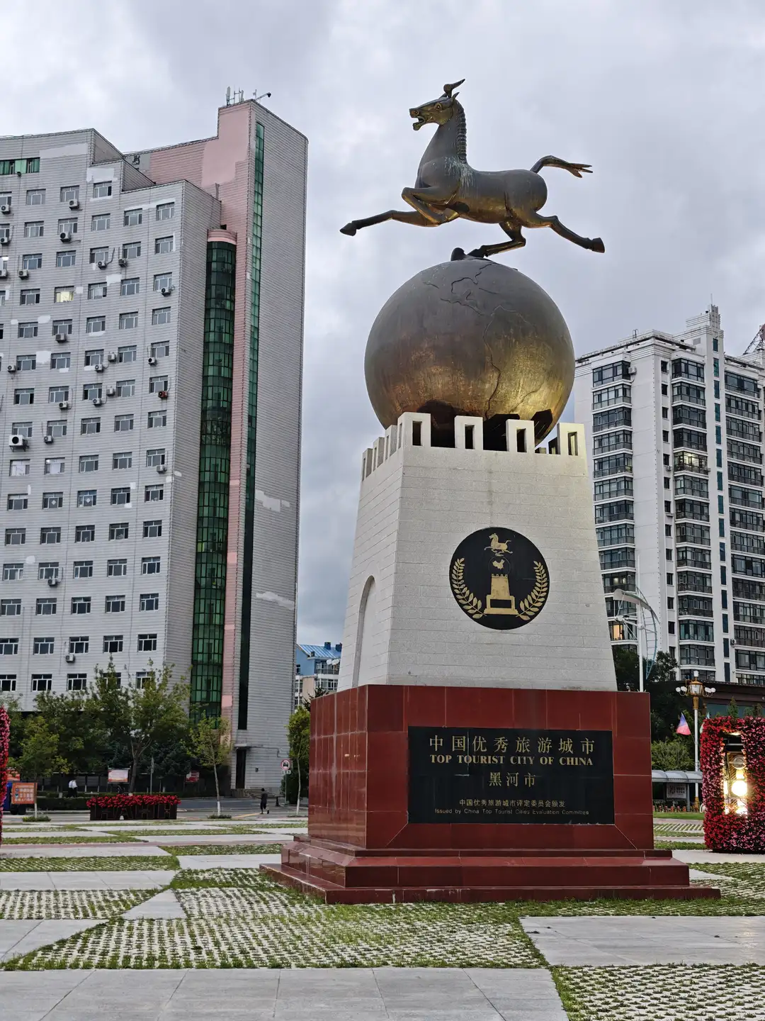 一座金色奔马雕塑矗立在一个球形基座上，球体置于方形石柱之上，石柱正面刻有“中国优秀旅游城市 黑河市”的字样。雕塑周围是铺有方砖的广场，背景是几栋高层建筑。基座底部为红色石材，整体结构显得庄重而醒目。