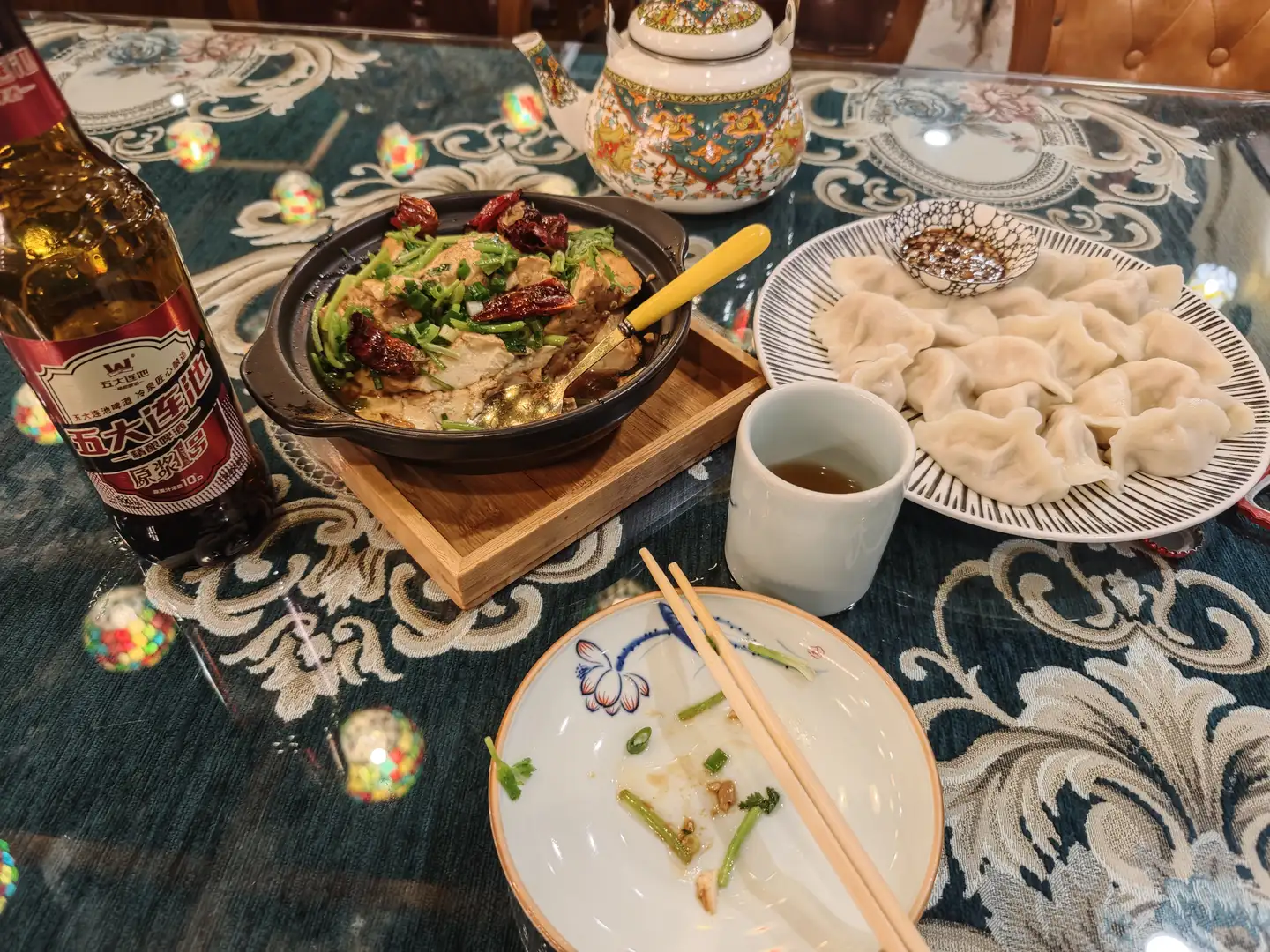 餐桌上摆放着一盘饺子、一锅热菜、一瓶啤酒和一杯茶。热菜盛在黑色砂锅中，上面有青椒和干辣椒，旁边放着一把黄色手柄的勺子。桌布是深绿色带花纹的，中央有一只空盘子，上面留有食物残渣和一双筷子。