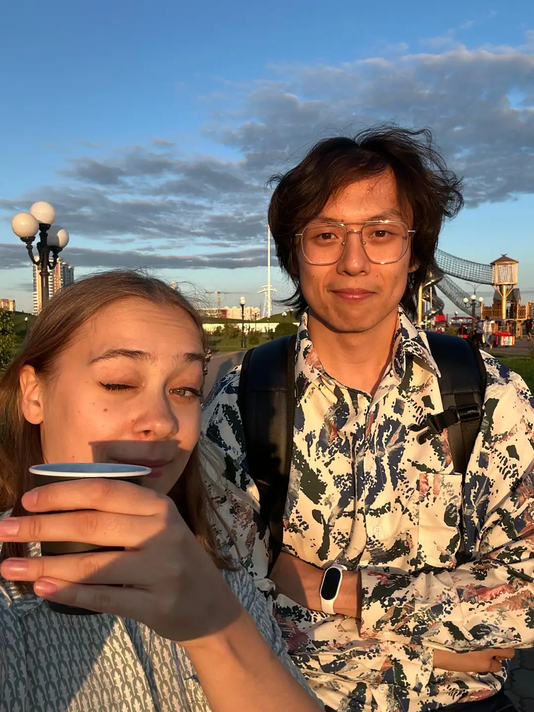 两个人在户外合影，其中一人手持杯子正在喝东西，背景有灯光柱和游乐设施。天空呈现蓝色并带有云彩，远处可见建筑物。