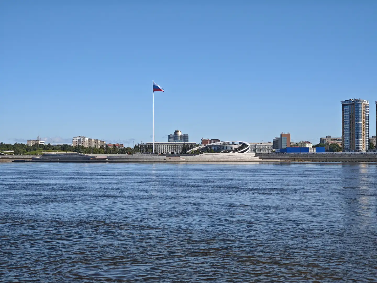 宽阔的水面延伸至对岸，岸边有高耸的旗杆上飘扬着俄罗斯国旗，周围是现代建筑群和一个独特的白色拱形结构。
