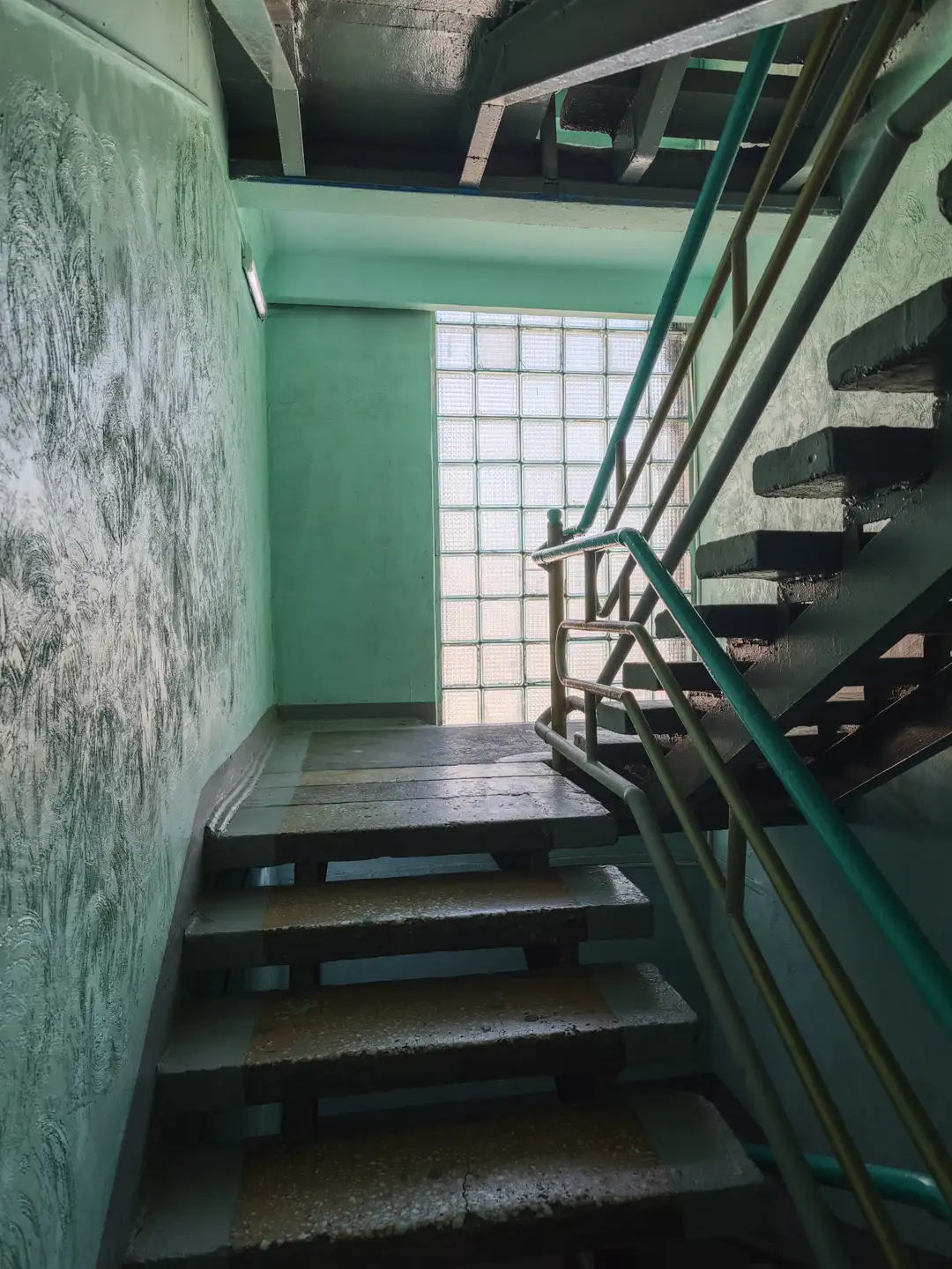 楼梯间墙壁呈绿色，表面斑驳，台阶由混凝土制成，边缘有黄色防滑条，右侧有金属扶手，尽头是一扇由玻璃砖组成的窗户。