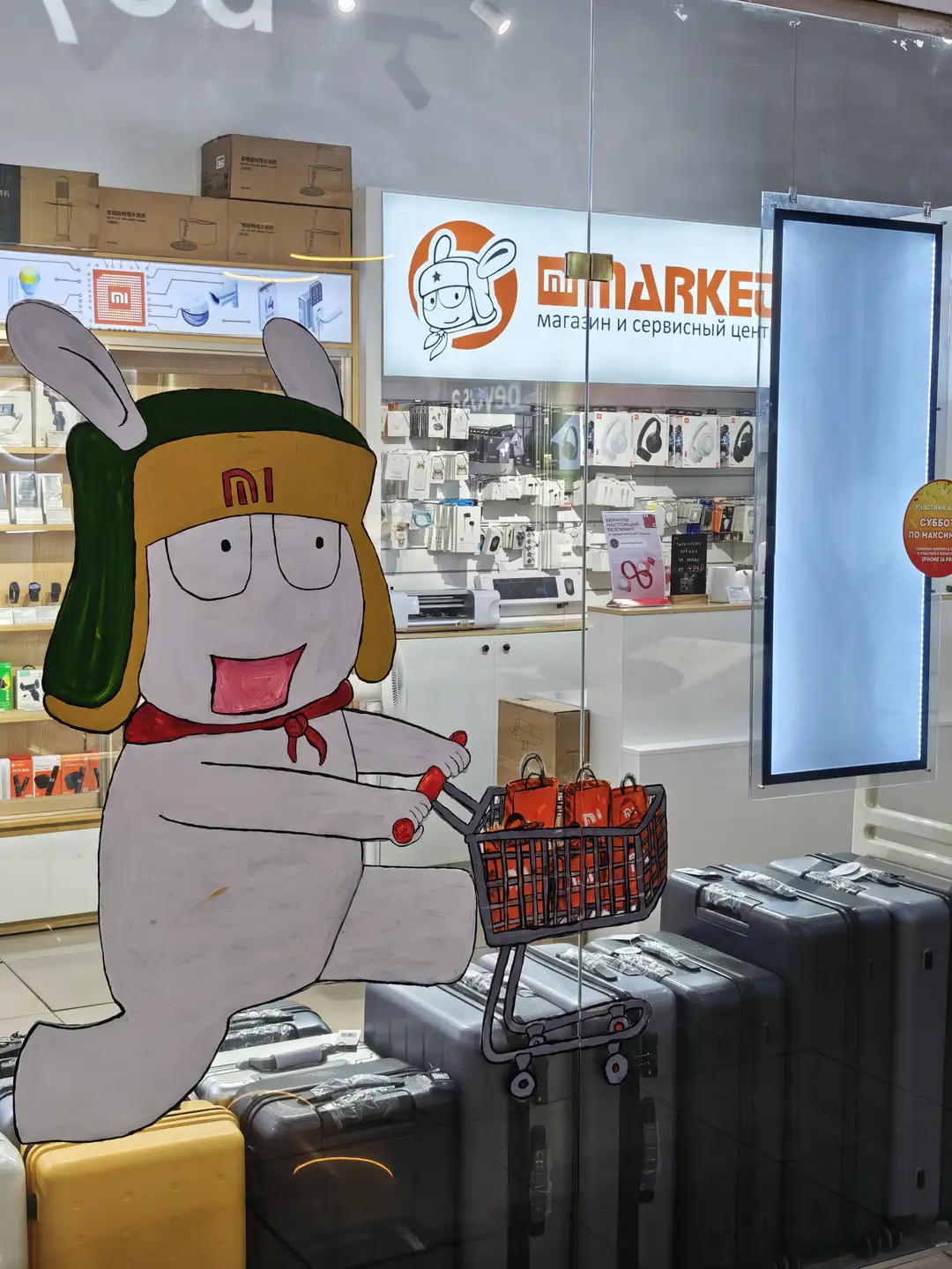 商店门口贴着一只卡通兔子形象，戴着印有“MI”标志的帽子，推着装满红色购物袋的手推车，店内陈列着电子产品和包装盒，招牌上写着“mi MARKET”。