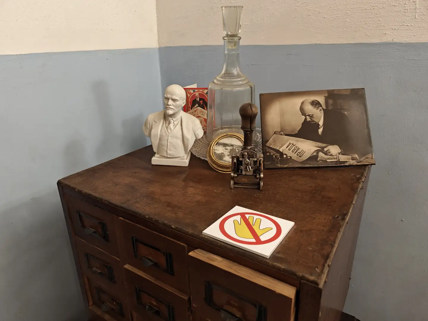 深色木柜上摆放着列宁半身像、玻璃瓶、老照片、印章和一个禁止触摸的标志，照片中人物正在阅读报纸，背景墙为浅蓝与白色拼接。