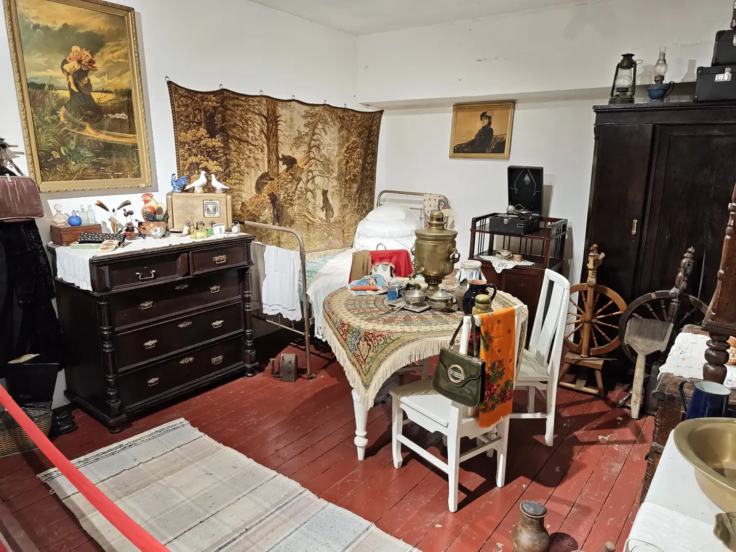 房间内陈设复古，有木质地板、铁床、衣柜和纺车，中央摆放着铺有花纹桌布的小圆桌，桌上放着茶具和铜壶，墙上挂着风景画和挂毯。